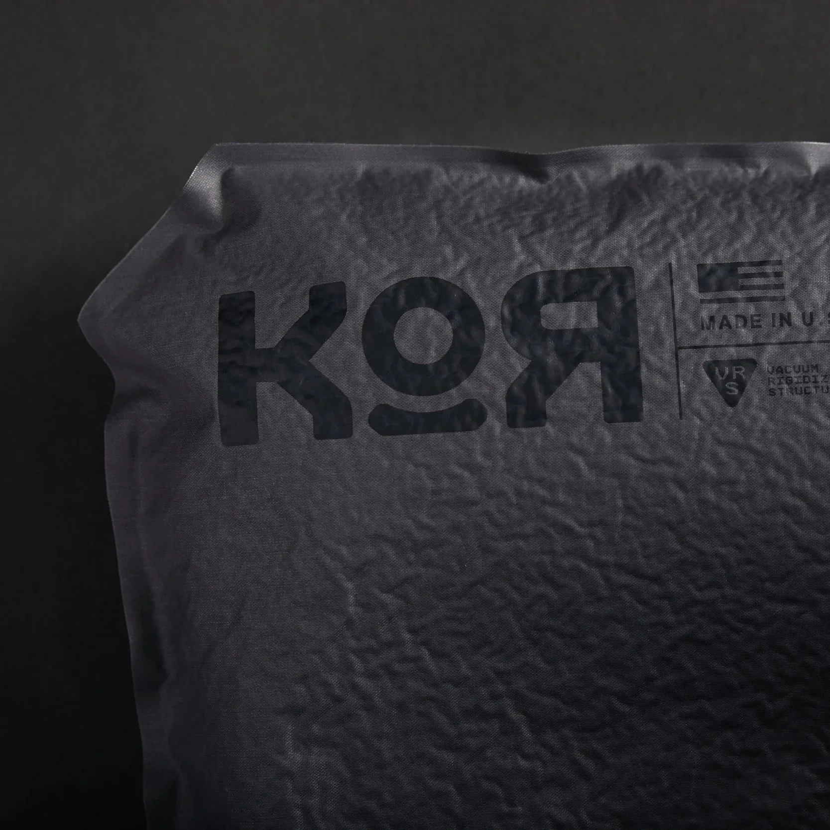 Close-up of a transparent vacuum-sealed plastic packaging with black text that says 'KOR' and includes recycling and manufacturing information, against a dark background.