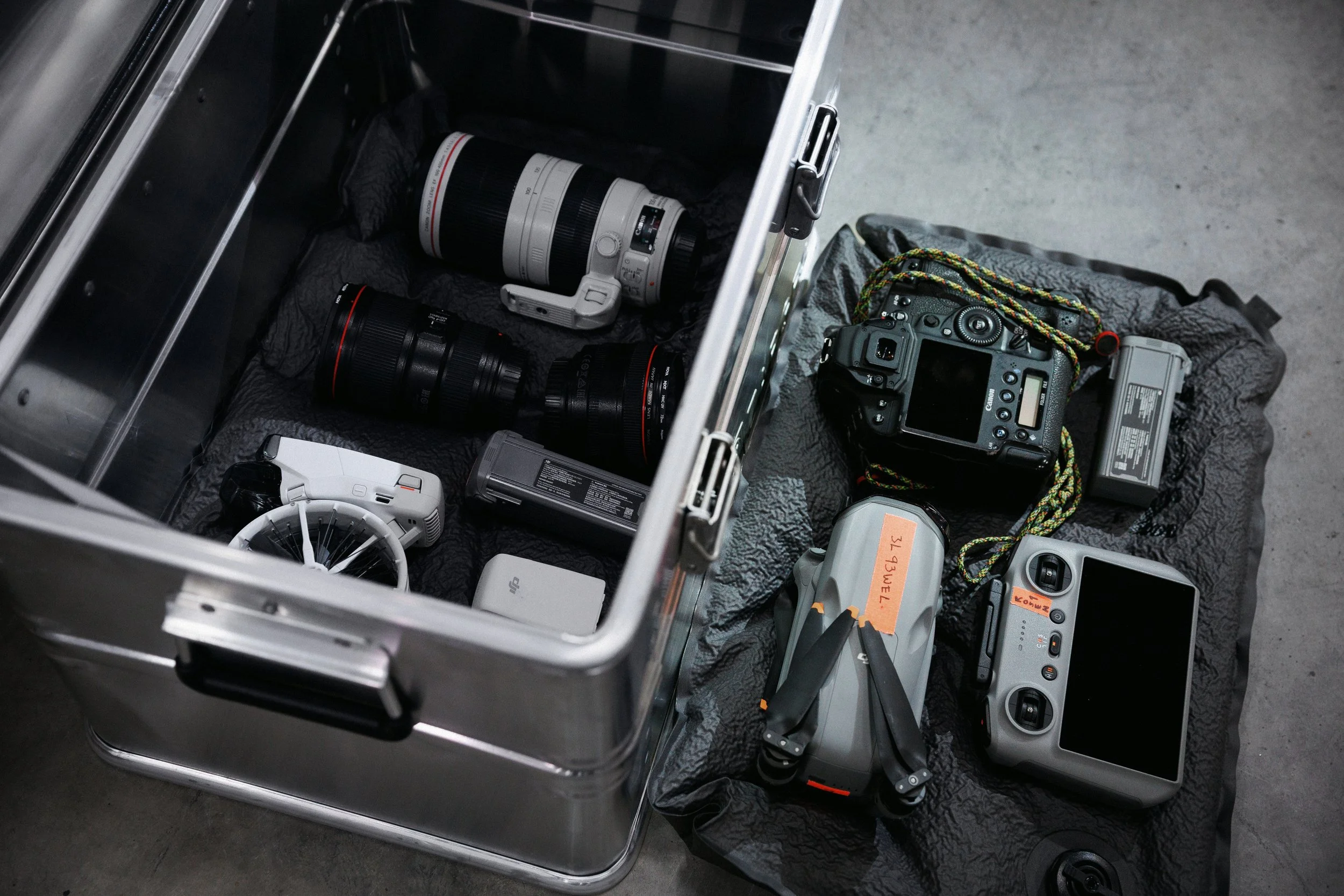 A metal equipment case containing camera lenses, a camera, and electronic devices, with some gear placed on a black cloth surface.
