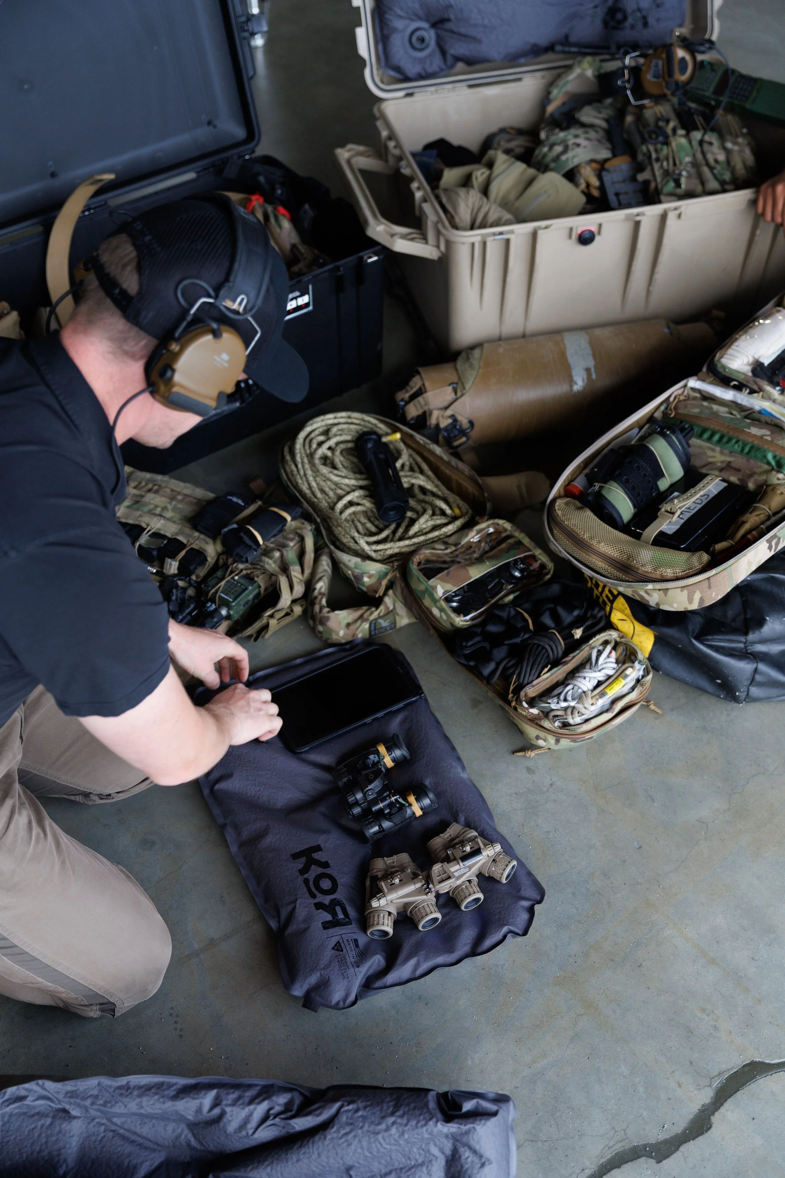 Person wearing headphones and a cap, kneeling on the floor, organizing tactical gear, including binoculars, ropes, and other equipment, with open cases and storage containers around.