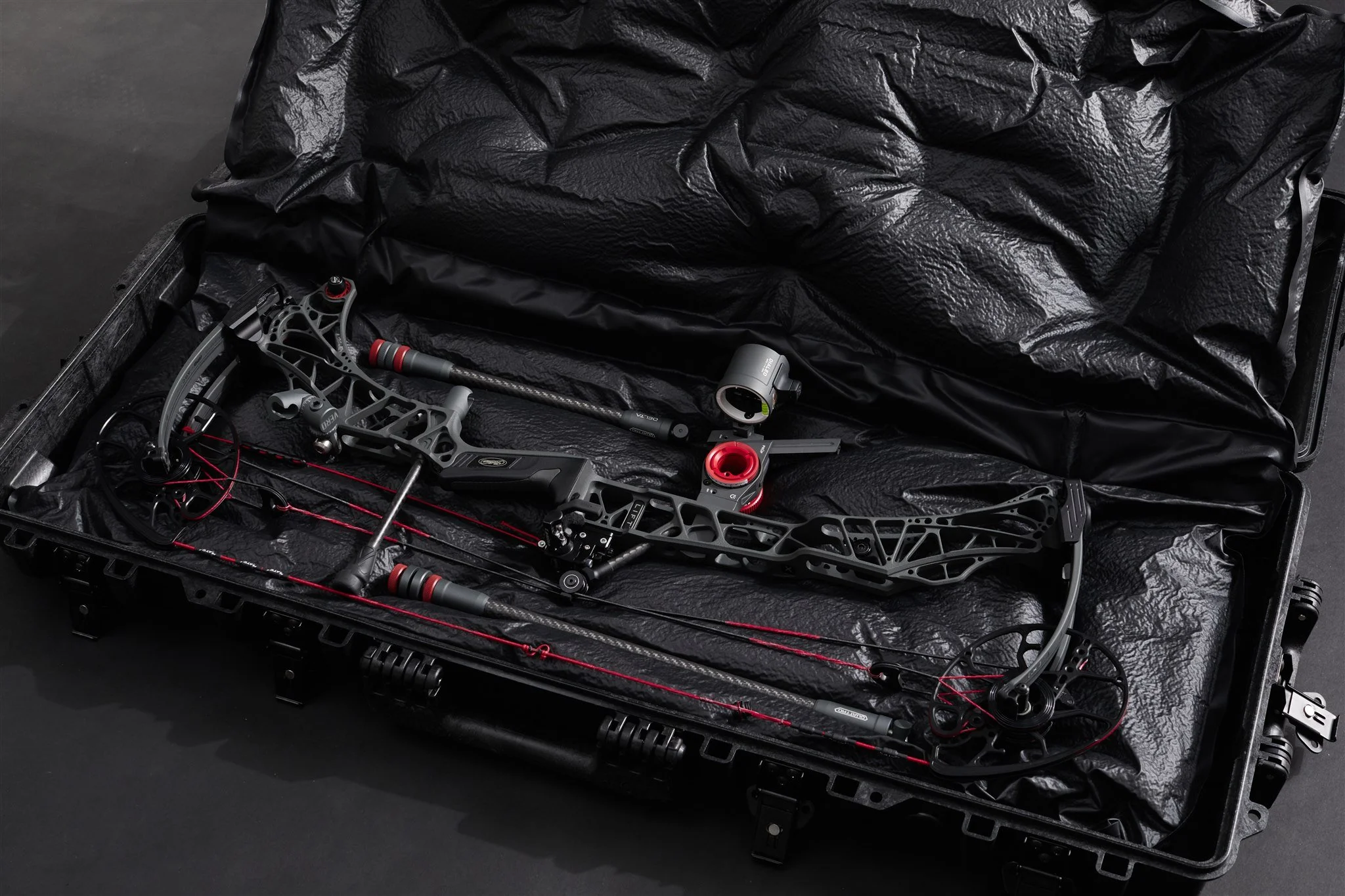 A black case with a crumpled black paper lining, containing a rifle scope, a bipod, and other firearm accessories.