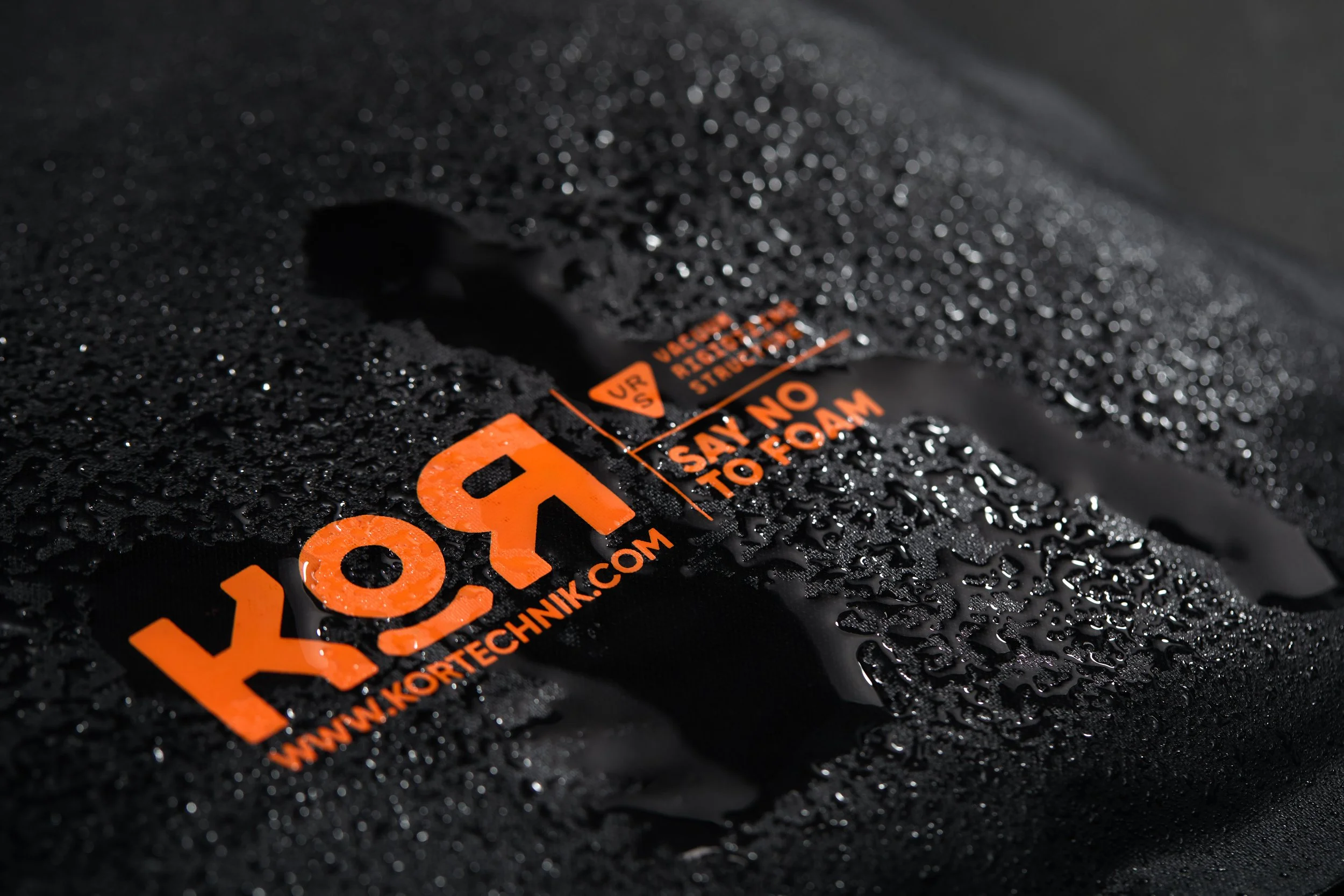 Close-up photo of a black, textured surface with water droplets. There is an orange label with black text partly visible on the surface.