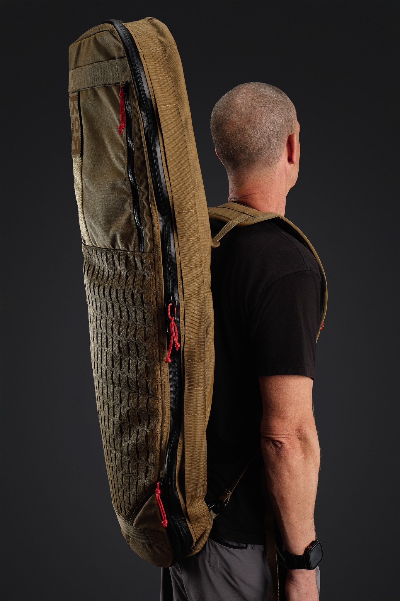 A person wearing a black t-shirt, gray shorts, and a smartwatch on their left wrist carrying a large, brown tactical backpack with multiple zippers and mesh pockets, standing against a plain black background.