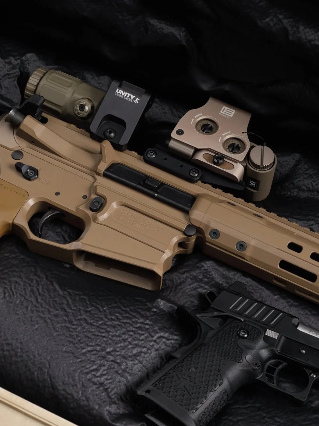 A tan-colored semi-automatic pistol, a tan rifle with attachments, and a black handgun are displayed on a black textured surface.
