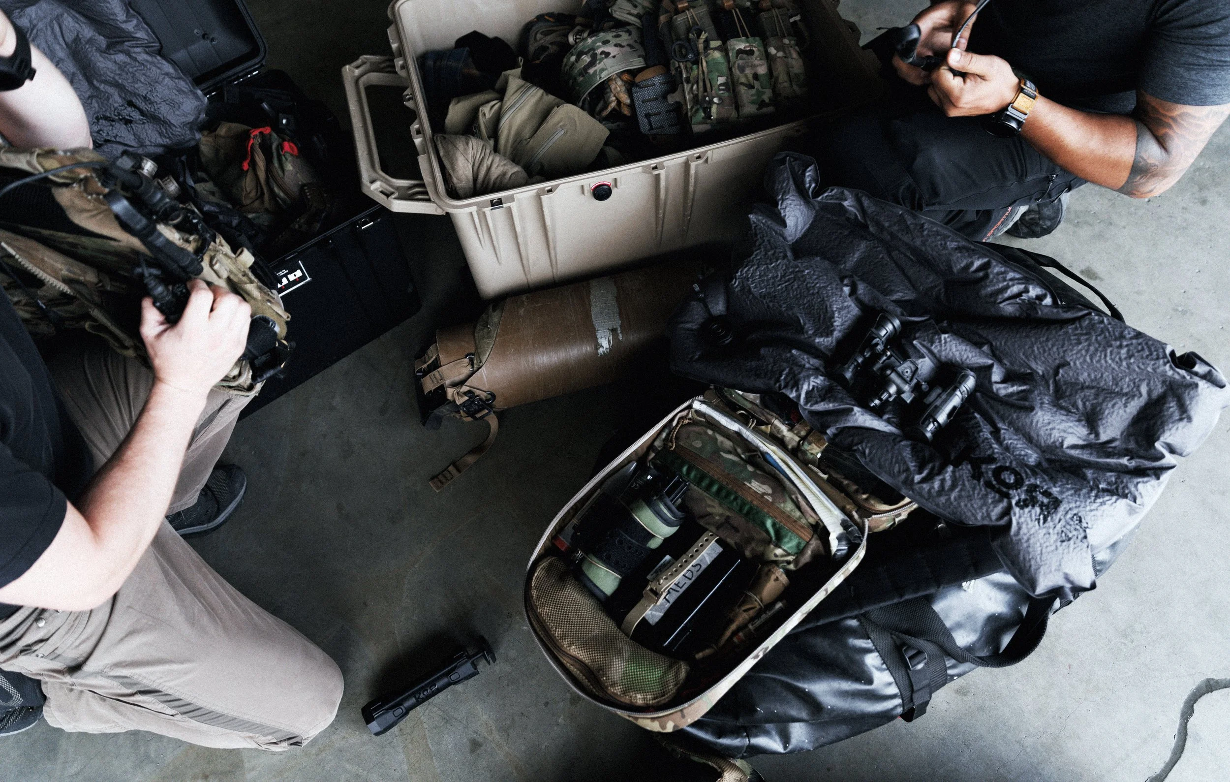 Open tactical gear and equipment bags with various military or outdoor gear, clothing, and binoculars on a table, with two people standing nearby, one wearing beige cargo pants and the other wearing black clothing and holding a headset with a microphone.
