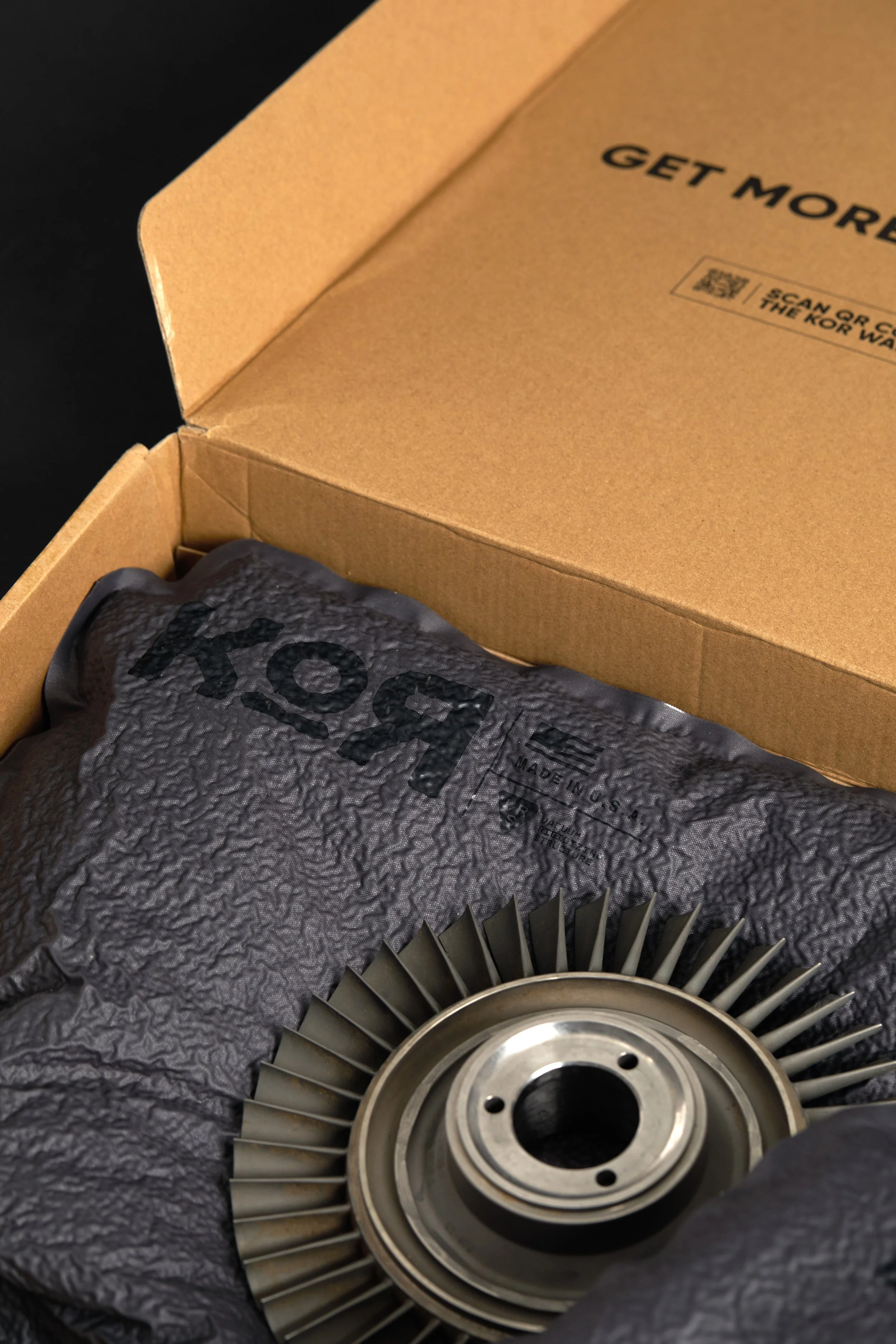 A metal turbine wheel resting on black textured padding inside a cardboard box. The box's top flap is partially open, revealing the turbine wheel.