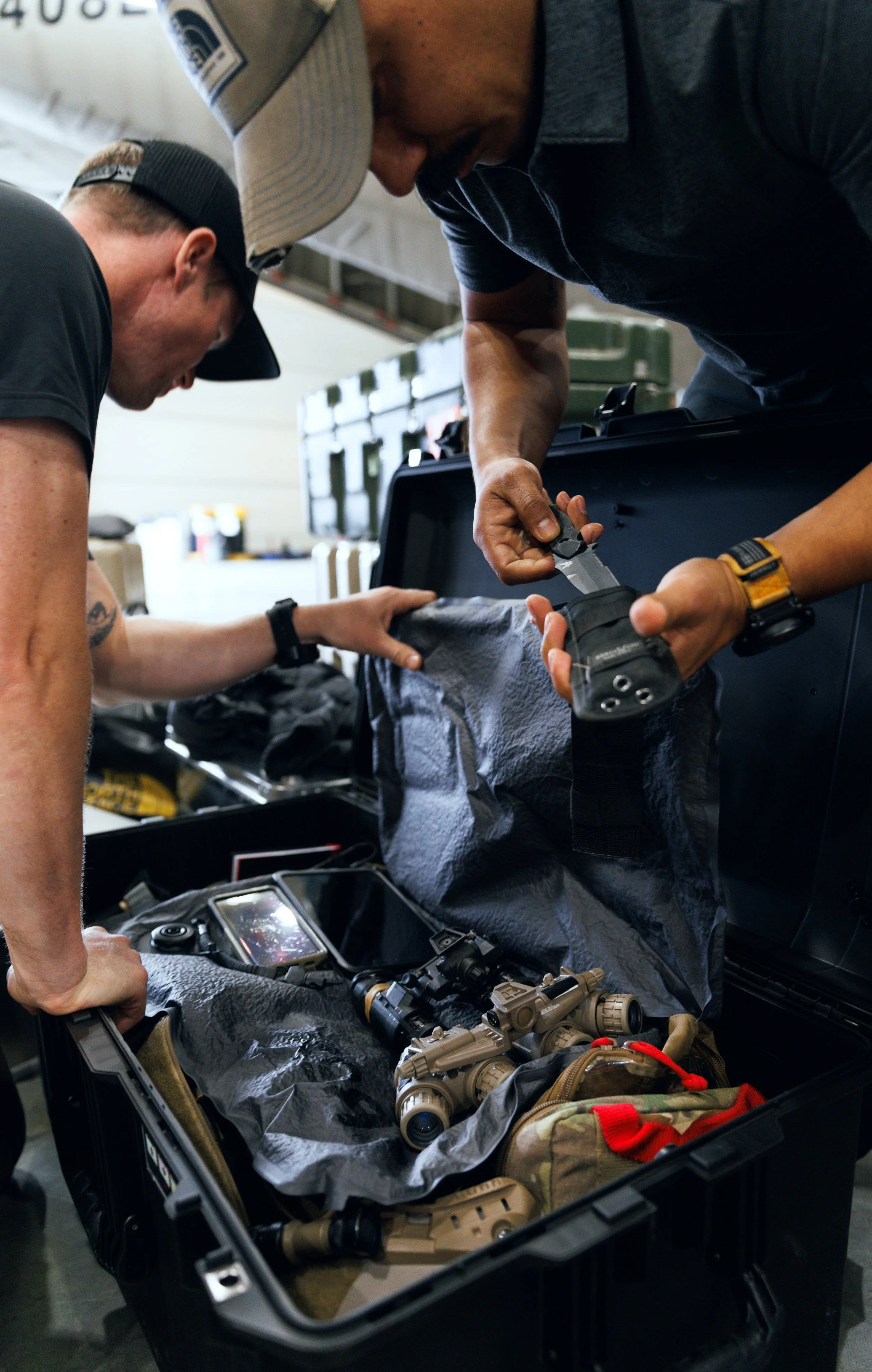 Two men organizing tactical gear, including scopes, a rifle, and other equipment, inside a black hard case.