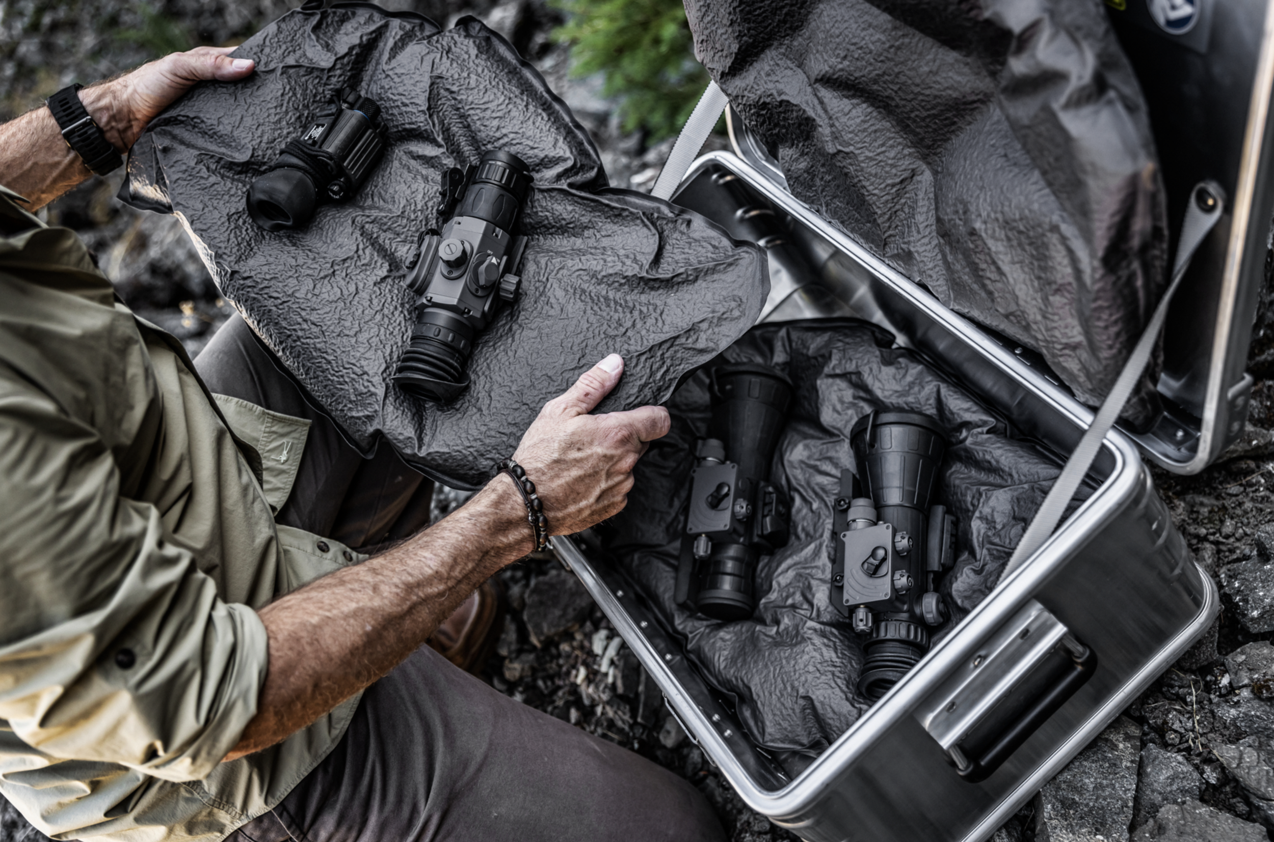 Person handling a black rifle with scope, placing it into a gray case with foam padding, outdoors on rocky terrain, wearing a beige jacket and beaded wristband.