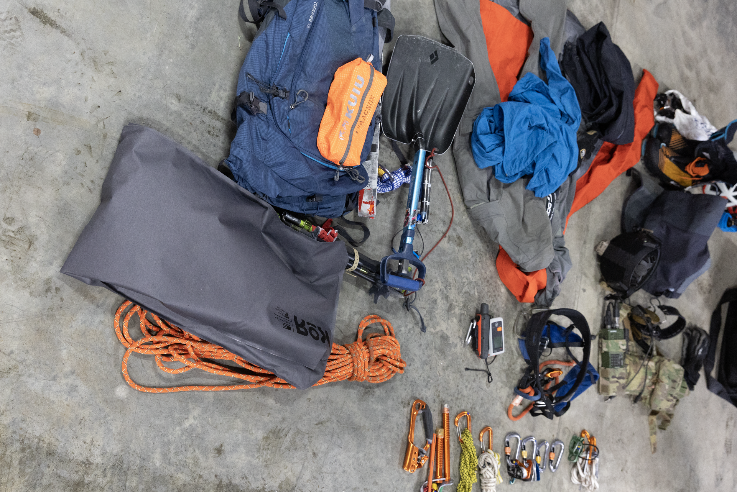 Climbing gear and equipment laid out on a concrete floor, including backpacks, ropes, carabiners, harnesses, and other safety accessories.
