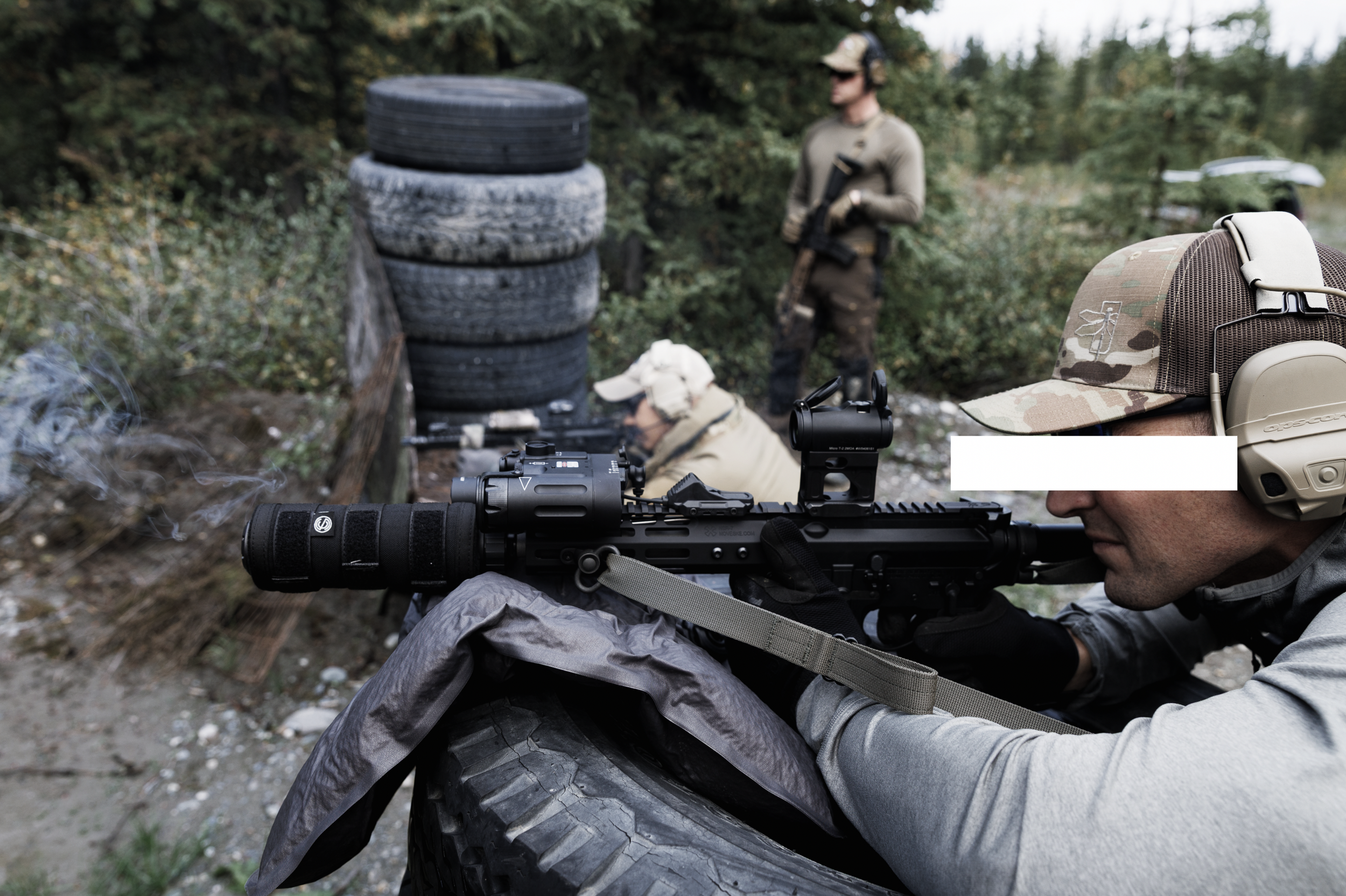A person aiming a rifle while lying on the ground outdoors with a scope attached, wearing a camouflage baseball cap and gray clothing, with a man standing in the background near a stack of tires, and a wooded area.