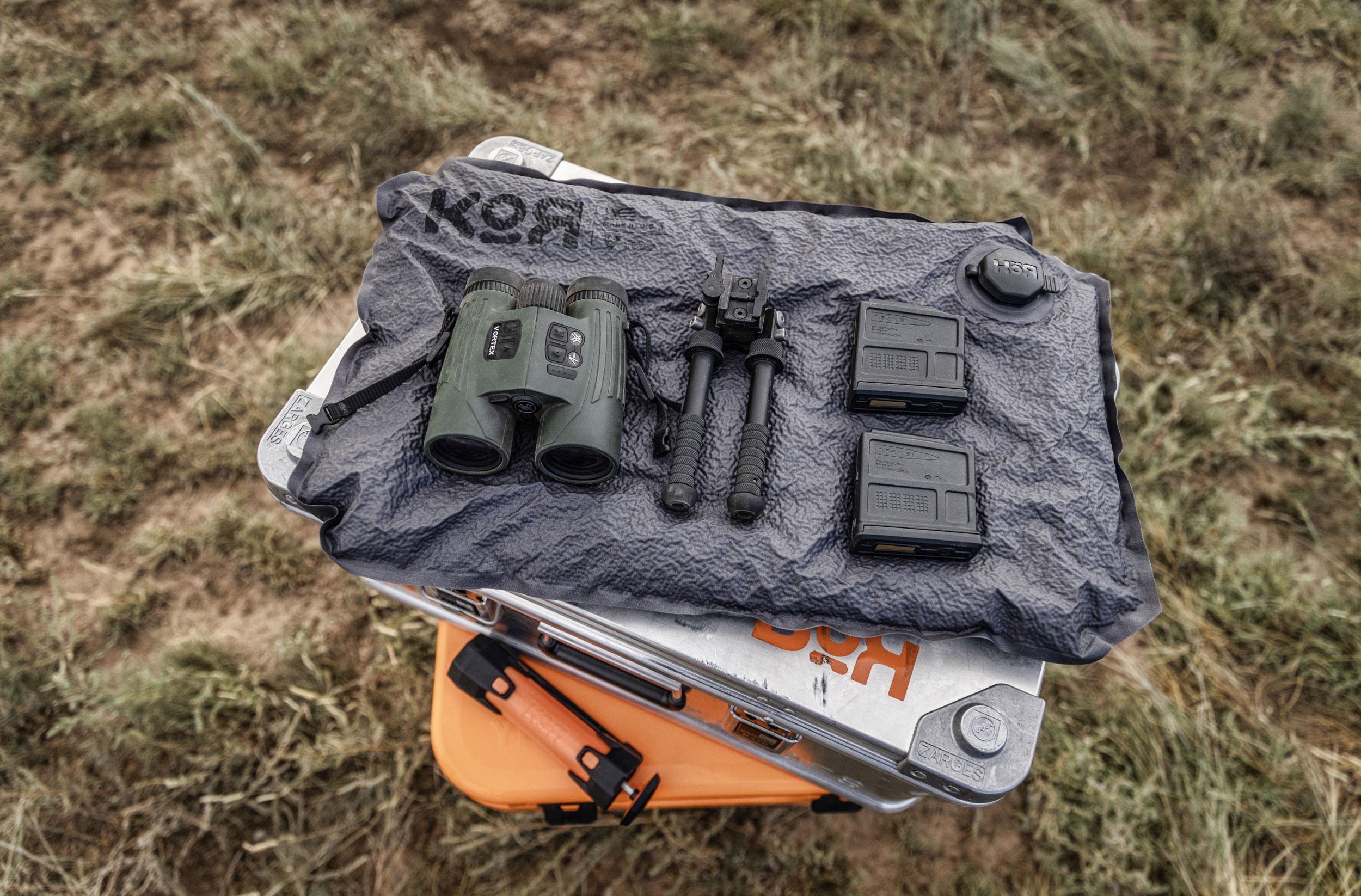 Camera and firearm accessories placed on a KOR panel on top of a Zarges case, outdoors on grass.