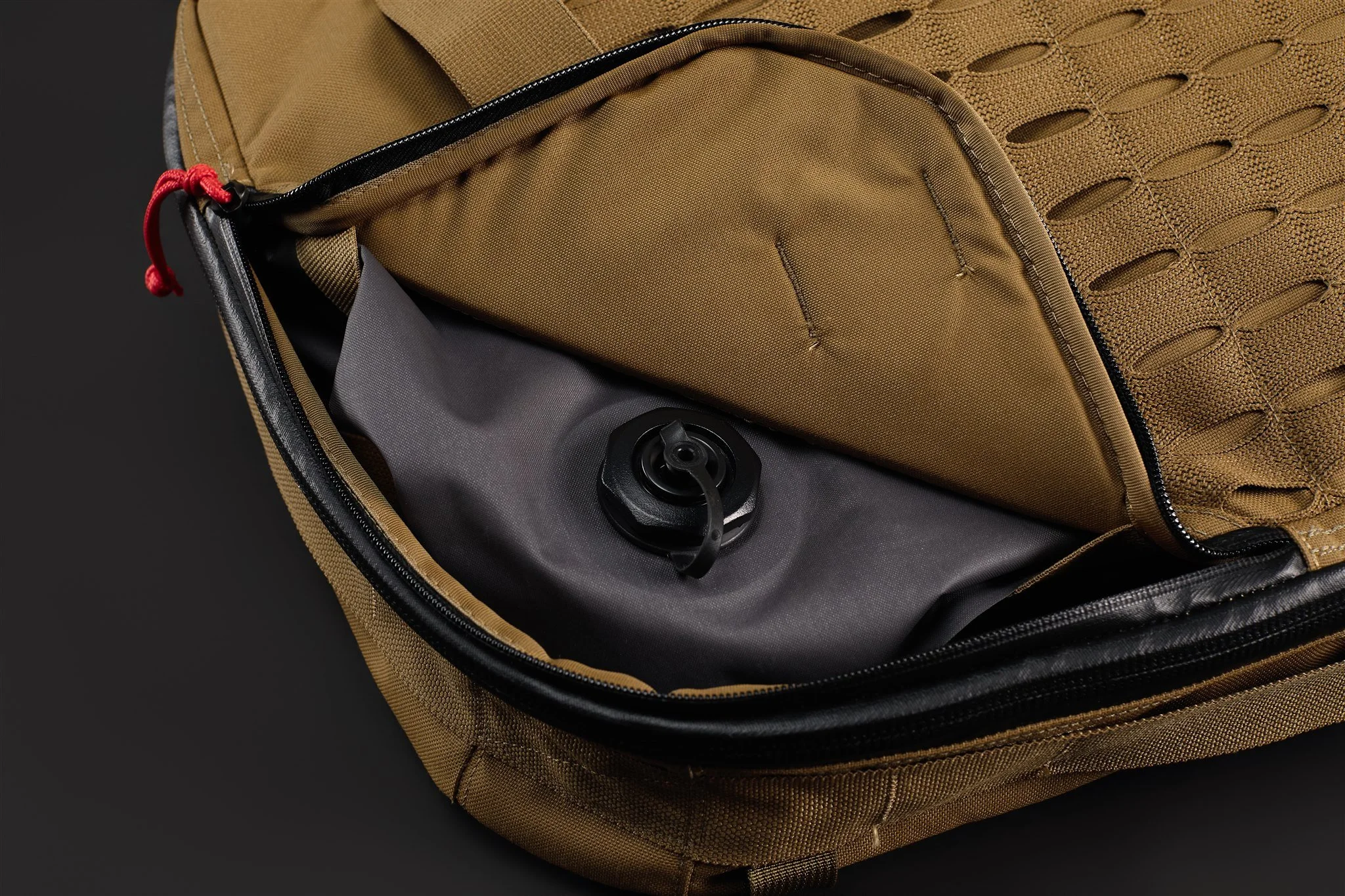 Close-up of a brown tactical backpack with an open zippered pocket containing a black object with a cord, against a black background.