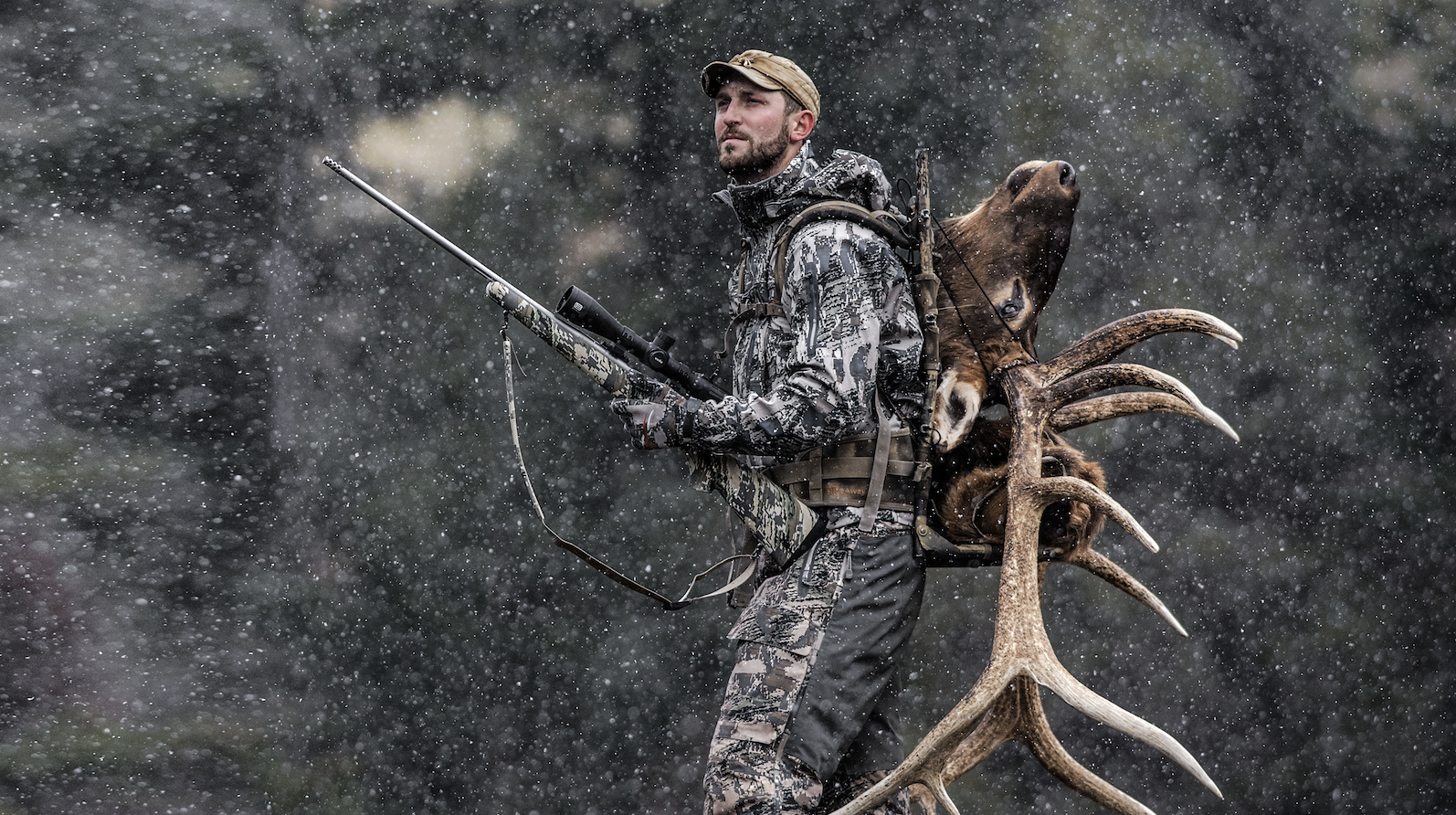A man dressed in camouflage hunting gear with a rifle, carrying a large set of elk antlers on his back, walking in a snow-covered forest.