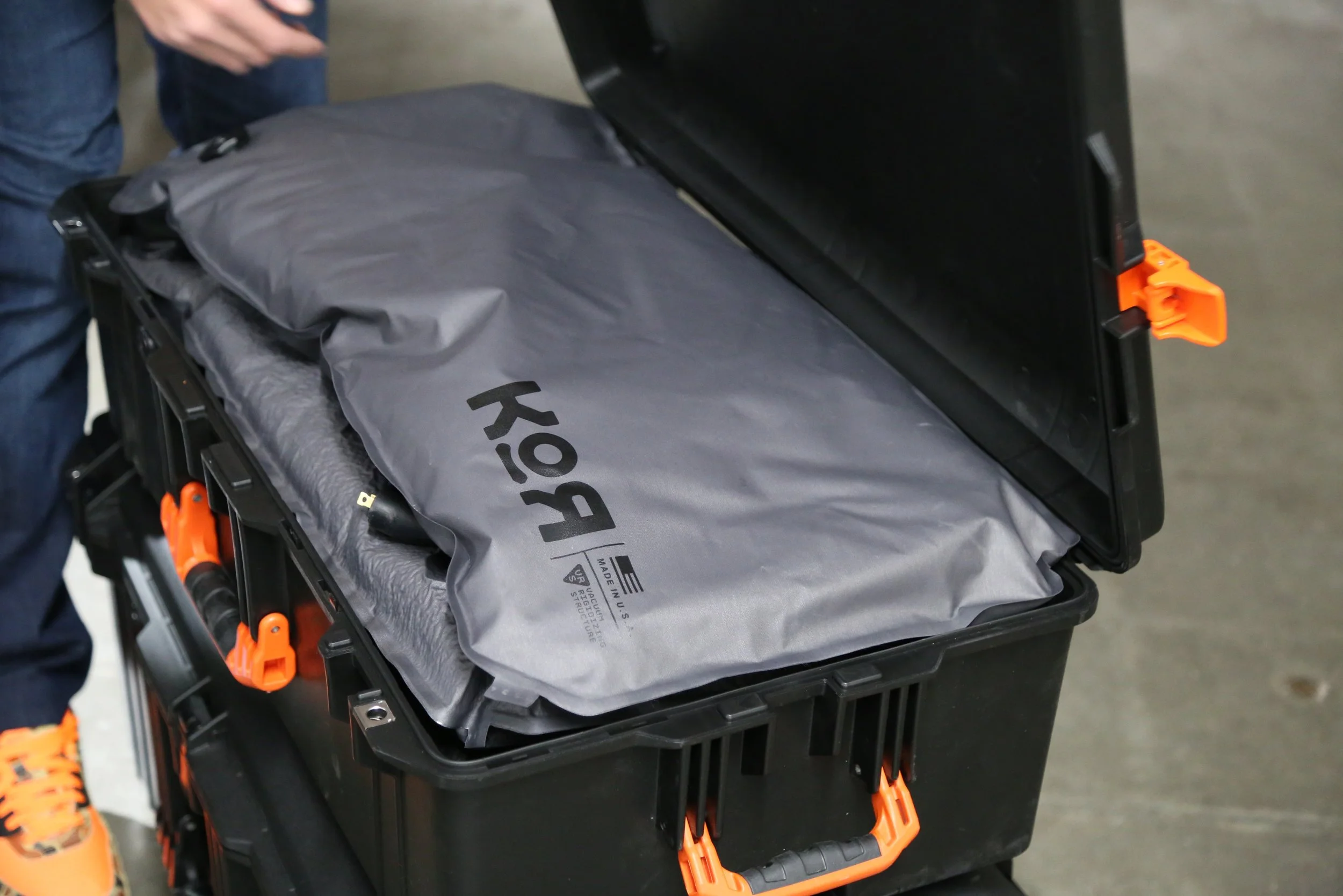 A black Pelican case, rolling cart with orange handles and clips, holding a KOR protection system panel KOR brand logo, is shown. A person in jeans and orange shoes is partially visible nearby.