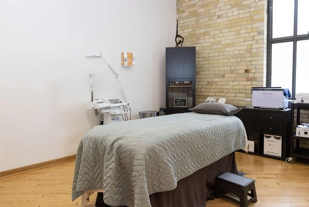 A medical examination room featuring a bed with a gray quilt, a pillow, and medical equipment, including a blood pressure cuff and monitor, against a backdrop of a brick wall and a large window.