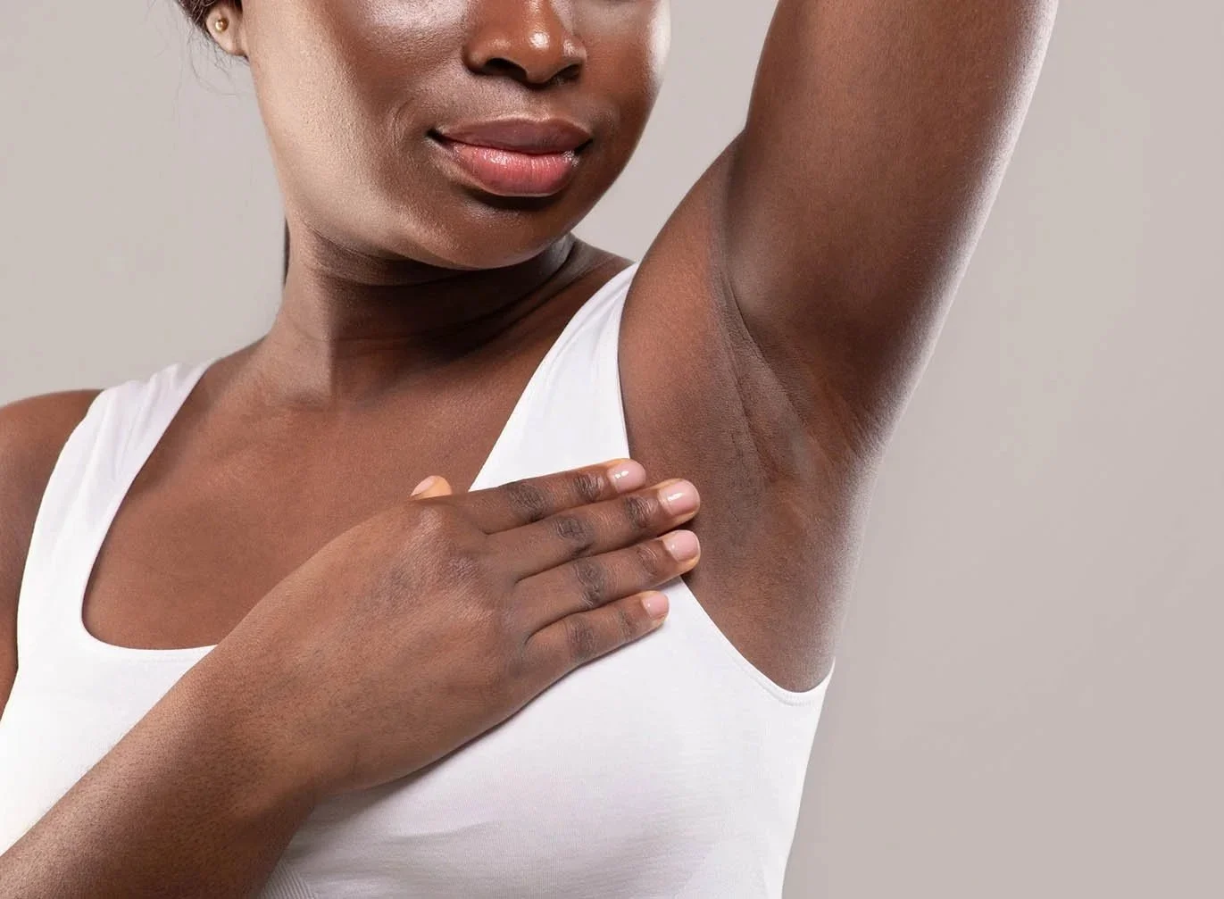 A woman with dark skin wearing a white tank top is lifting her arm to show her underarm, which is hairless and smooth.