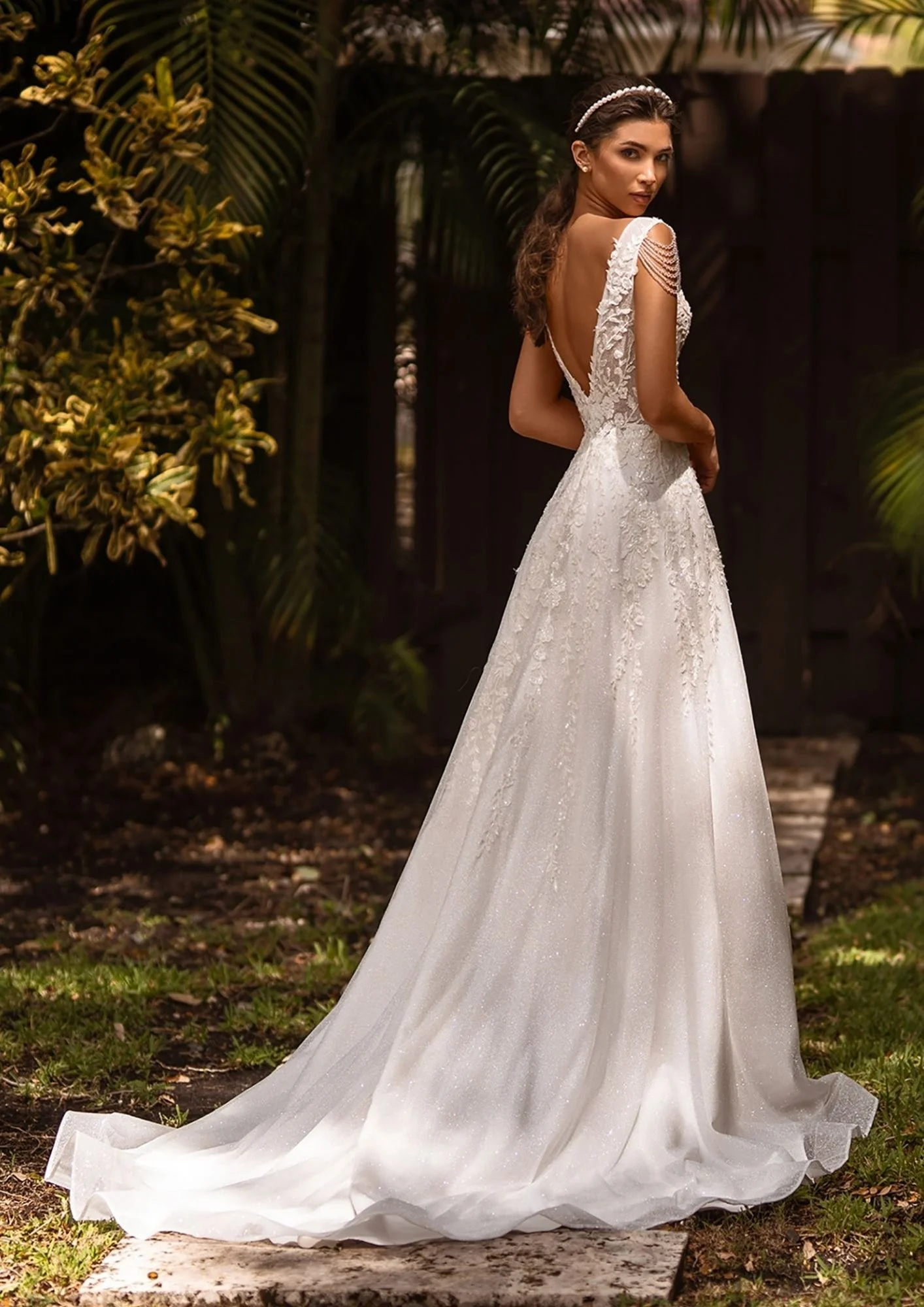 A woman in a white wedding dress standing outdoors among tropical plants, looking over her shoulder.