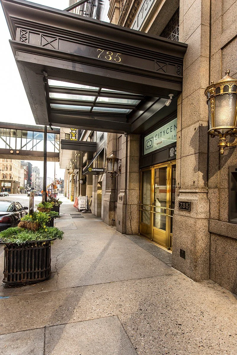 City street sidewalk with planter boxes, a building entrance with a glass door and brass frames, decorative lanterns, and a black canopy overhead, with the number 735 displayed above.