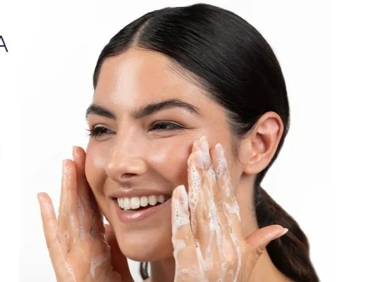 A woman with dark hair smiling as she applies soap to her face for skincare.