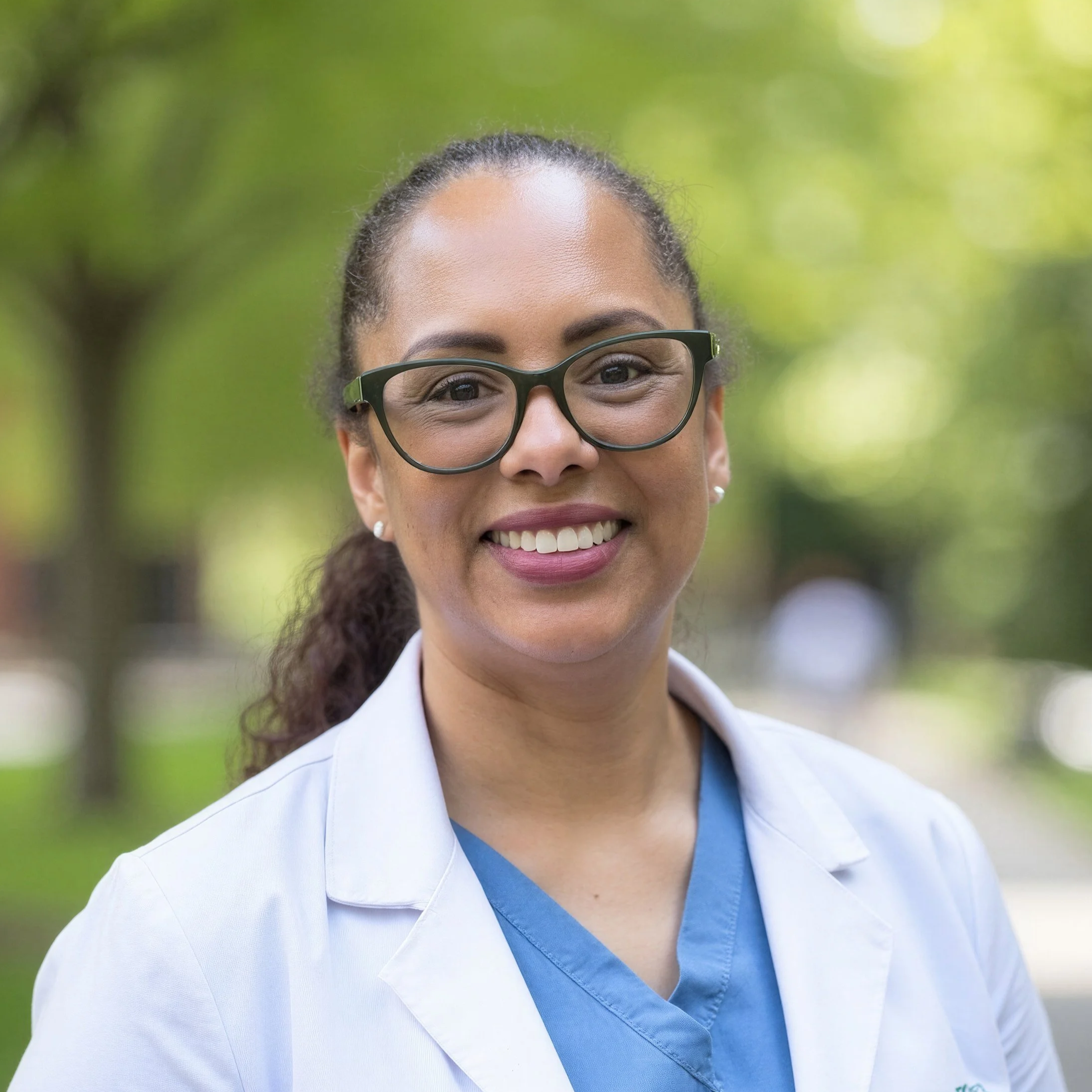 A woman with dark hair pulled back, wearing glasses, a white lab coat, and scrubs, smiling outdoors with a background of green trees.