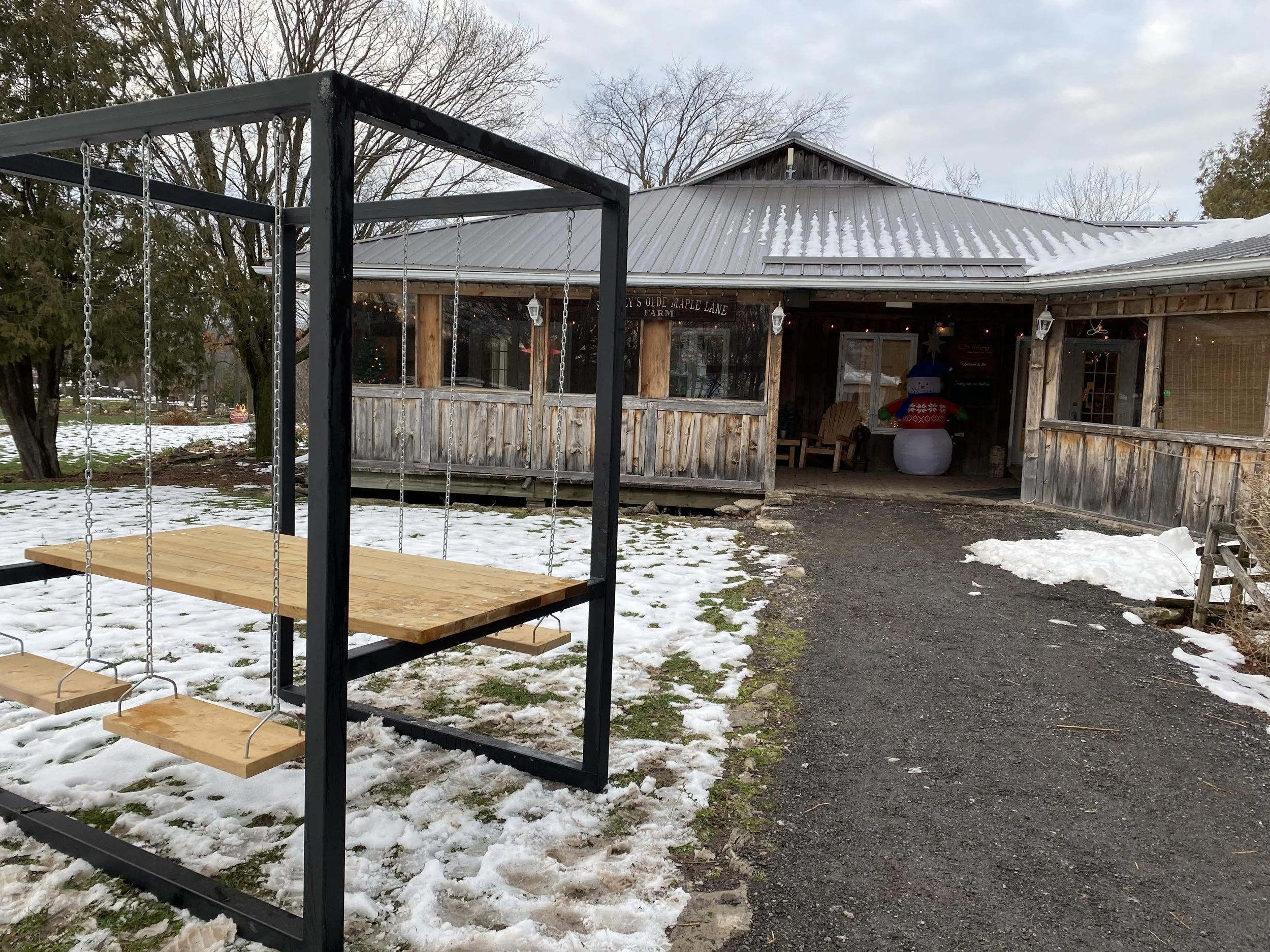 A rustic wooden farm building with festive holiday decorations, including a snowman, decorated porch, and snow-covered ground, with a custom steel swing set in the foreground.