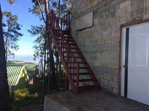 Red metal staircase leading to an upper level outside a stone building, with a door beside it, overlooking trees and a blue sky.