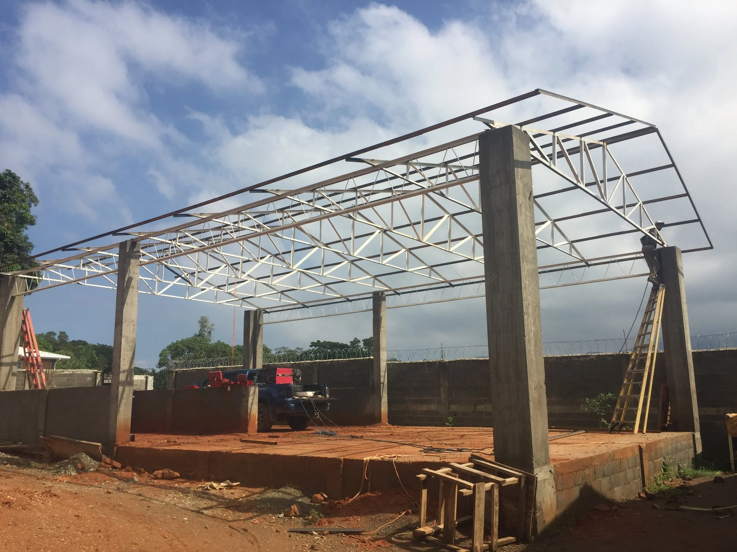 Construction site with a steel frame structure being assembled on concrete pillars, with work tools and ladders nearby, under a partly cloudy sky.