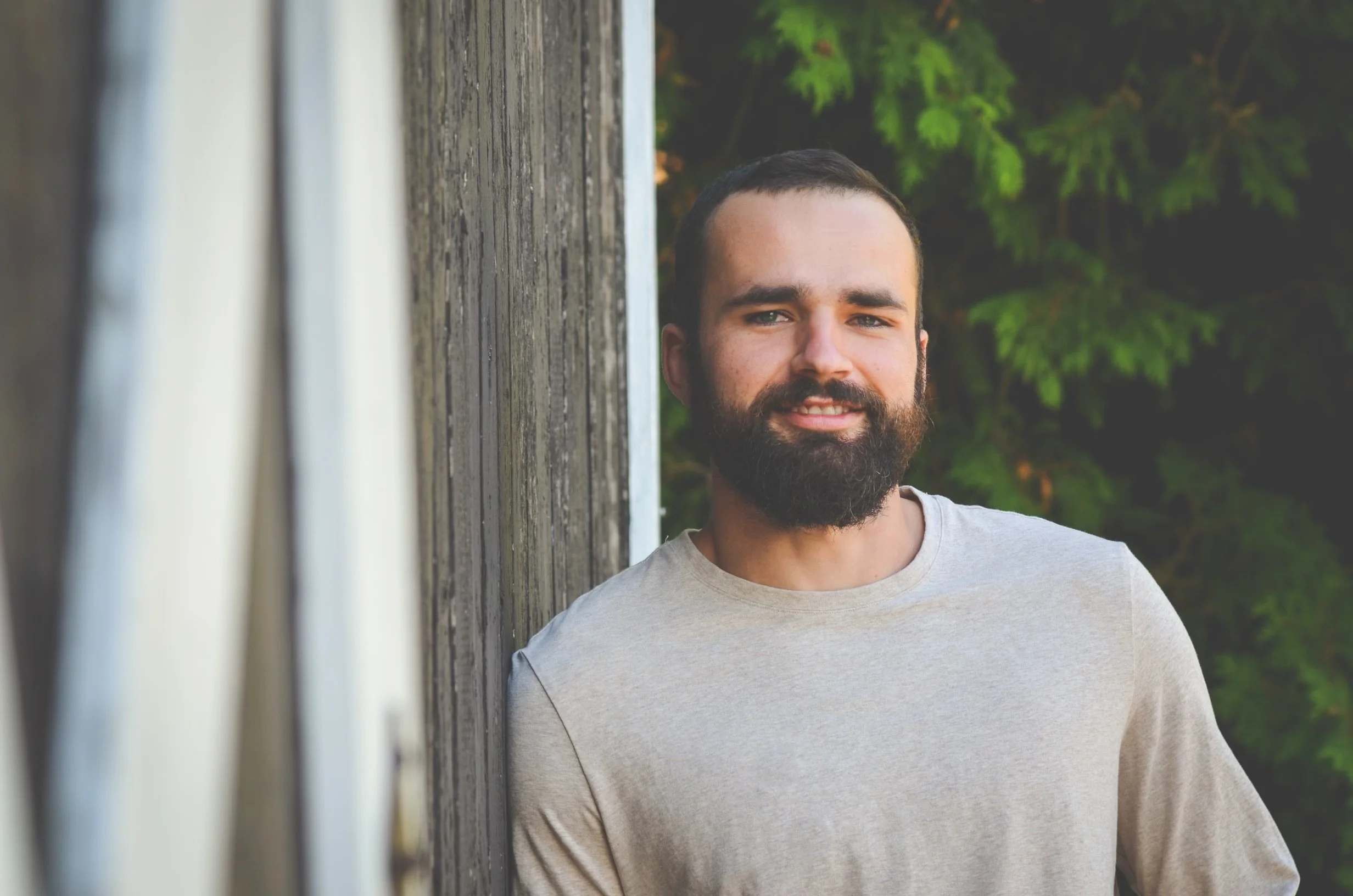 A man with a beard and short hair standing outdoors, leaning against a wooden wall, with green foliage in the background.