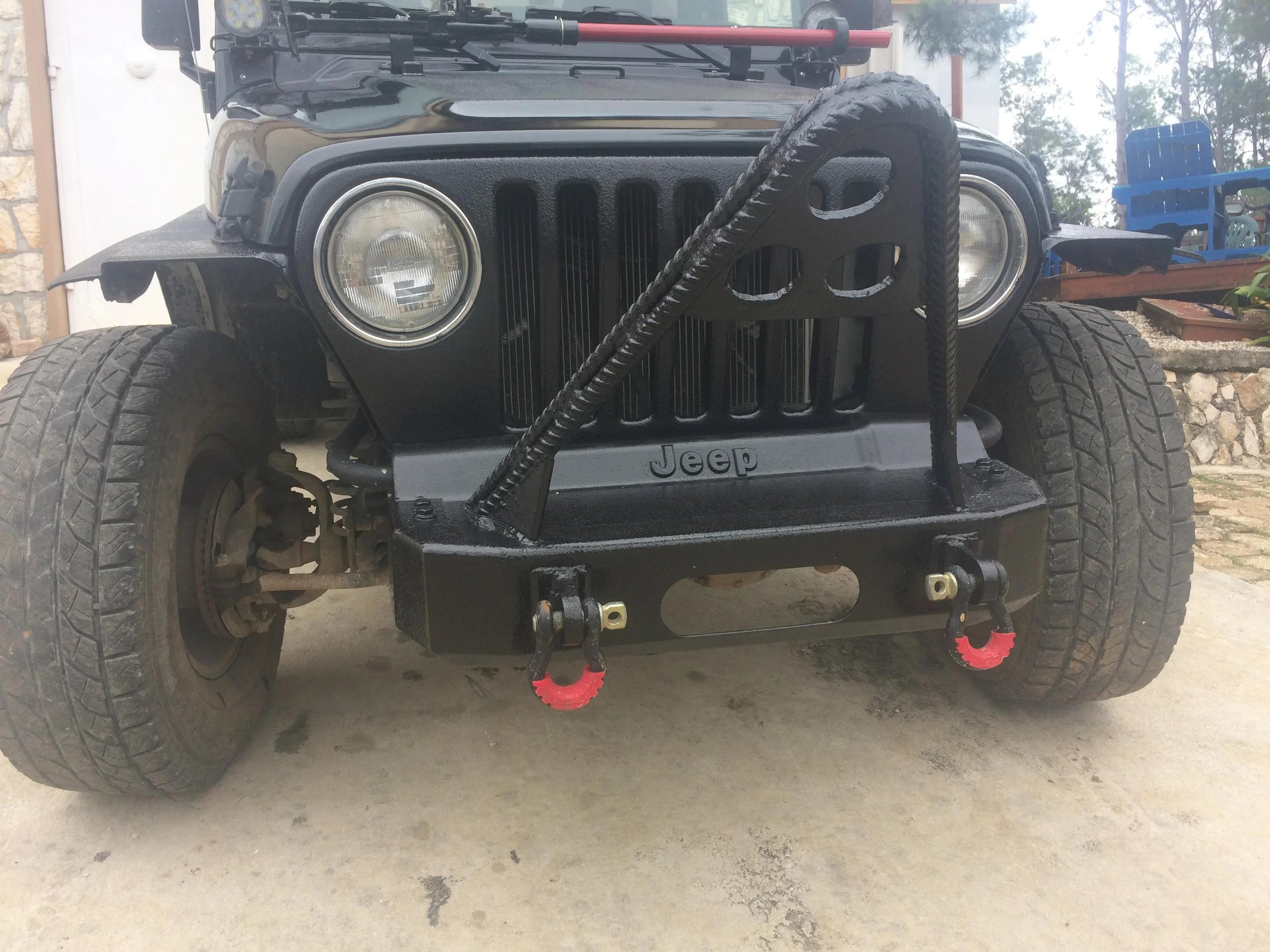 Close-up of the front of a black Jeep vehicle with large off-road tires, a custom fabricated bumper with red tow hooks, and a winch hook attached to the bumper.
