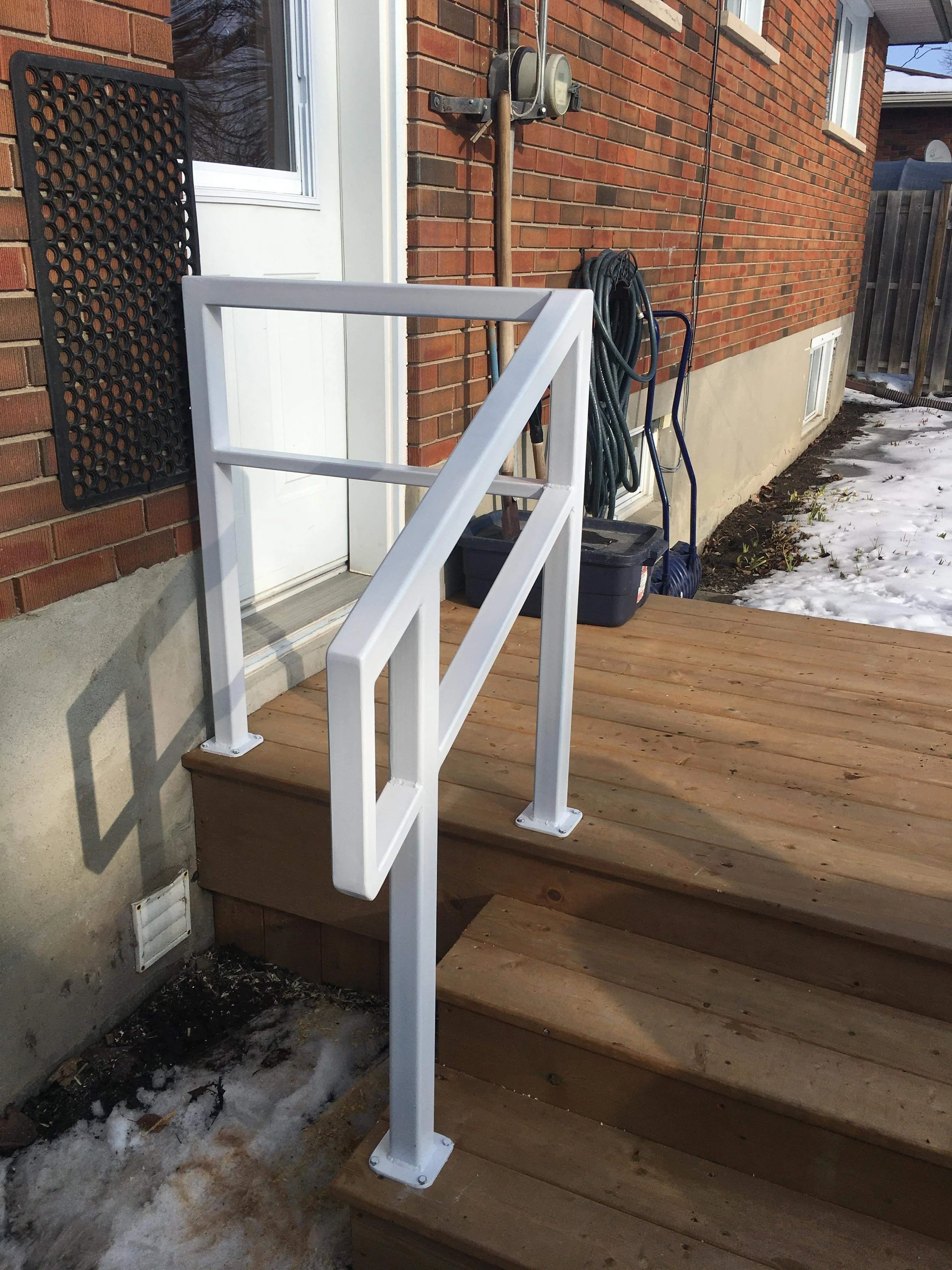 A white handrail installed on a wooden porch at a door entrance, with a brick house exterior and gardening tools in the background.