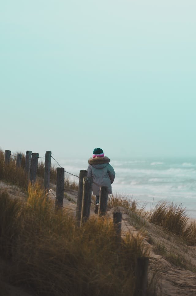 person in coat on a dirt path by a beach