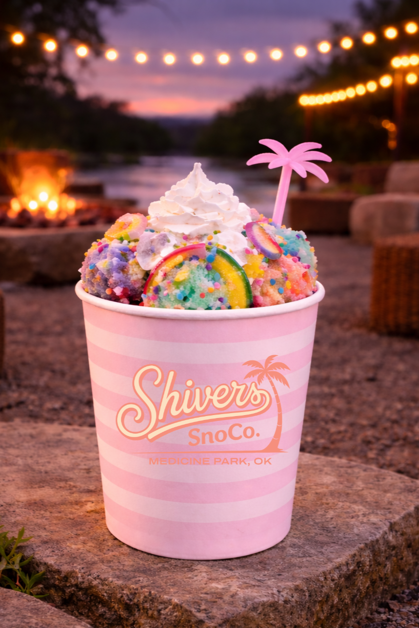 Cup of colorful ice cream with rainbow sprinkles, whipped cream, and a pink palm tree-shaped spoon, outdoors at sunset with string lights and a fire pit in the background.