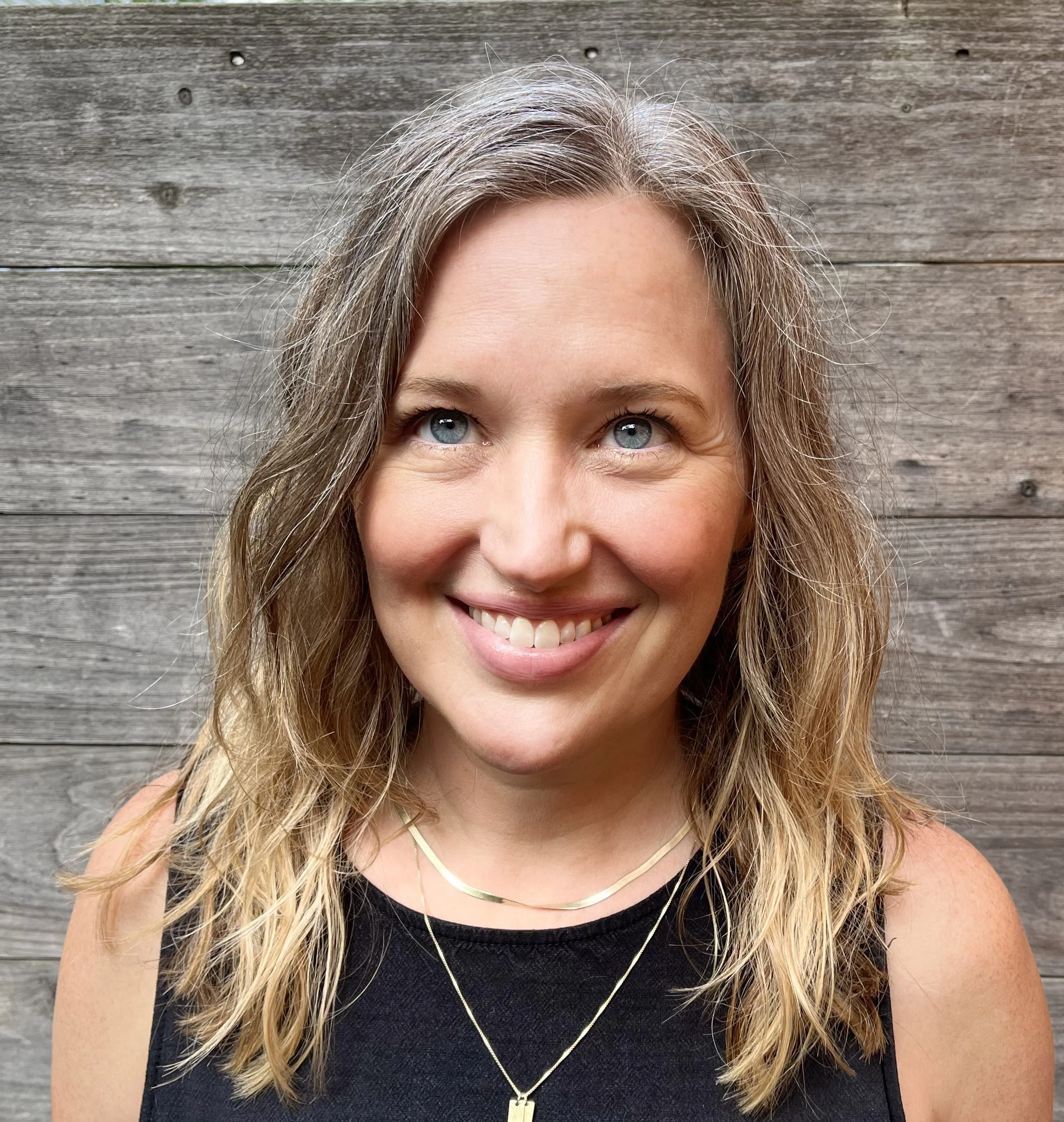 Close-up of a smiling woman with blue eyes and blond, wavy hair, wearing a black top and layered necklaces, standing in front of a wooden wall.