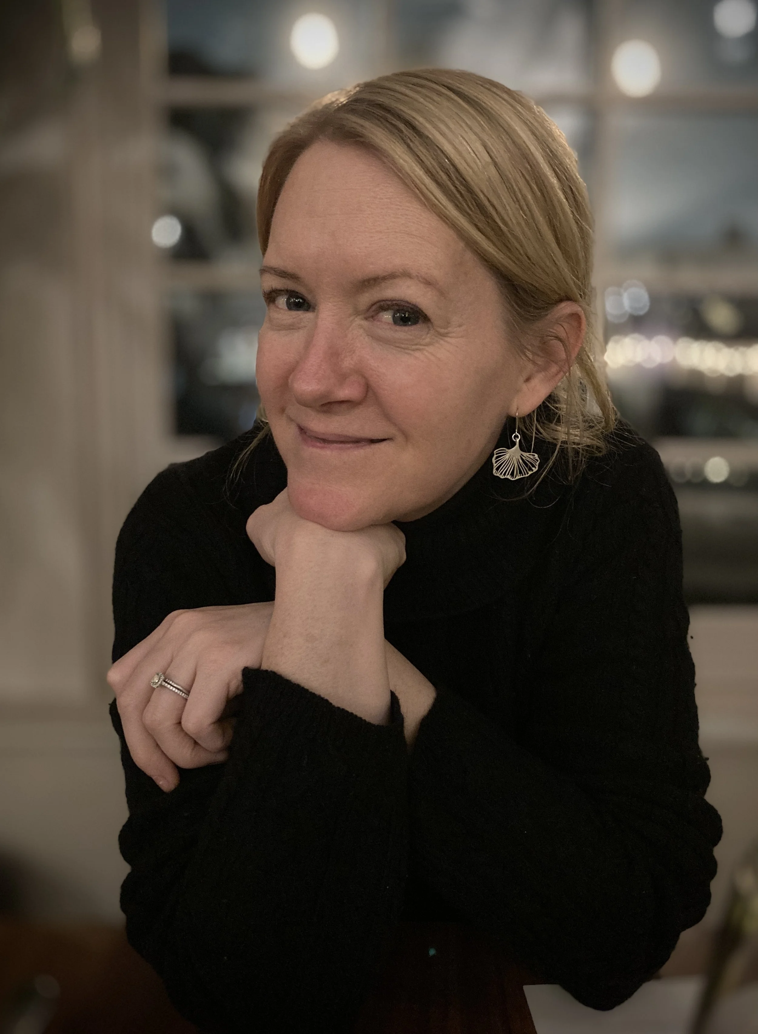 A woman with reddish hair wearing a black top, silver leaf-shaped earrings, and a ring on her finger, resting her chin on her hand and smiling at the camera. The background is blurred with soft bokeh lights.