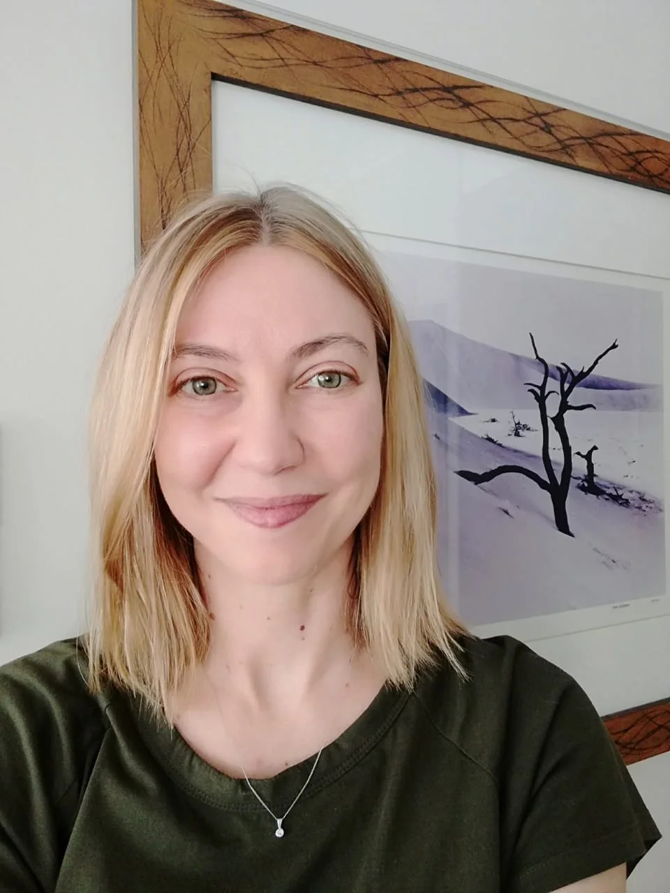 A woman with shoulder-length blonde hair smiling for a selfie, wearing a dark green top and a silver necklace, standing in front of a framed artwork of a snowy landscape with a barren tree.