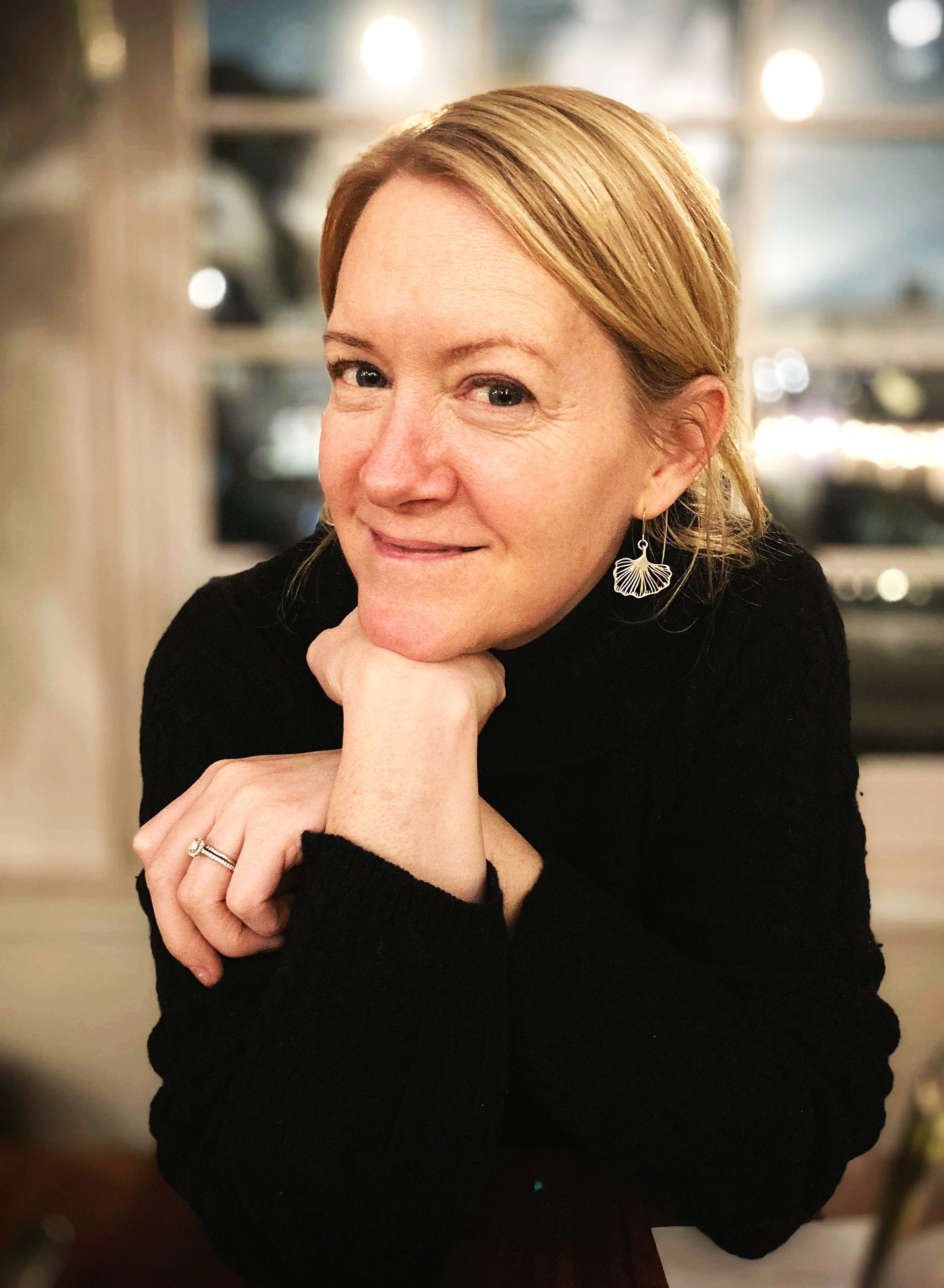 A woman with red hair smiling and resting her chin on her hand, wearing a black top and a hanging earring, in a cozy indoor setting with warm lighting.