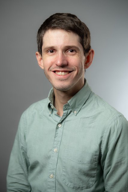 A smiling man in a light green collared shirt posing against a gray background.