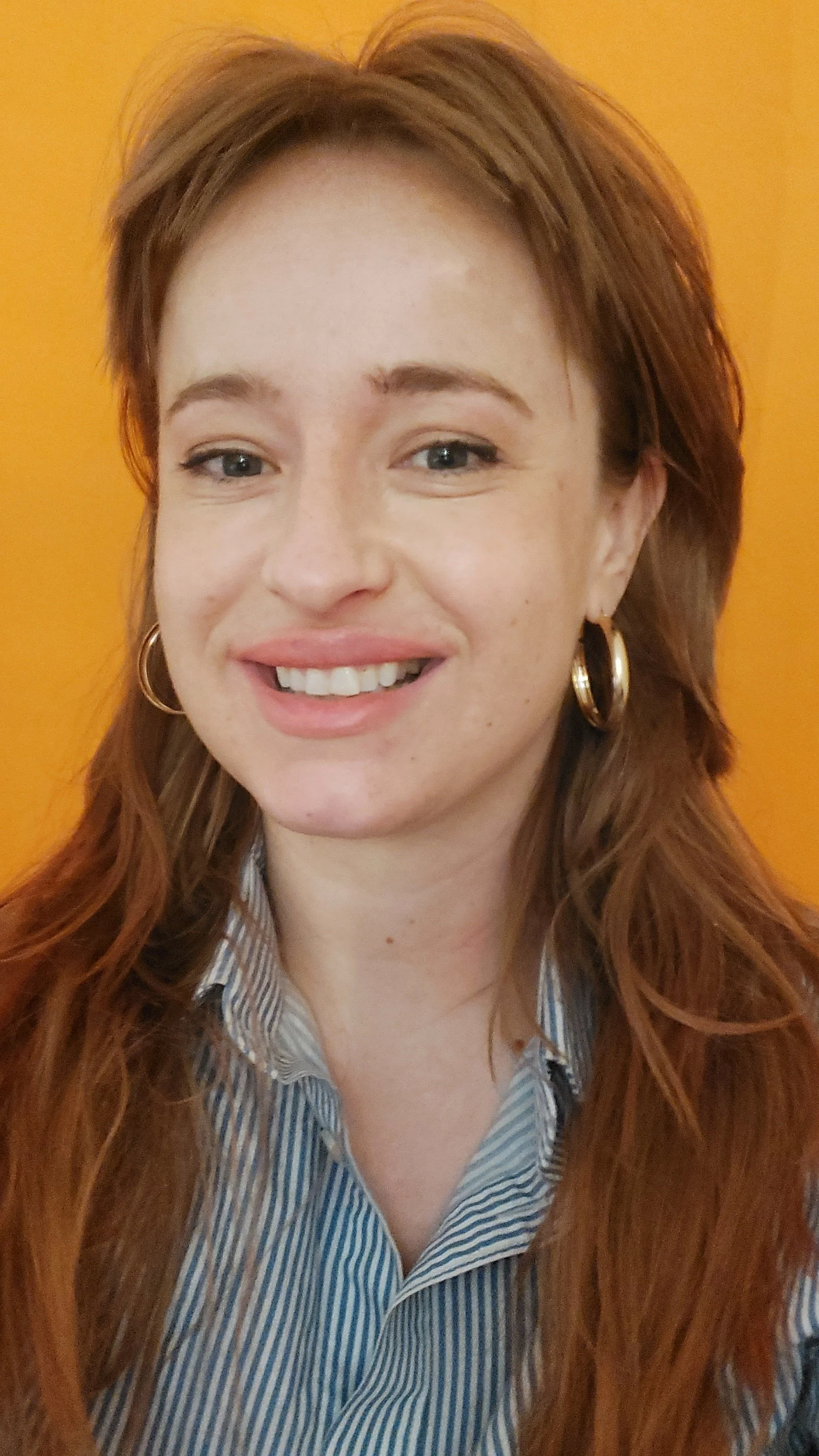 A woman with long, wavy red hair wearing a striped shirt, hoop earrings, and smiling against a yellow background.