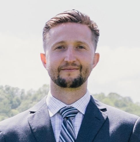 Headshot of a man in a suit and tie outdoors with a blurred natural background.
