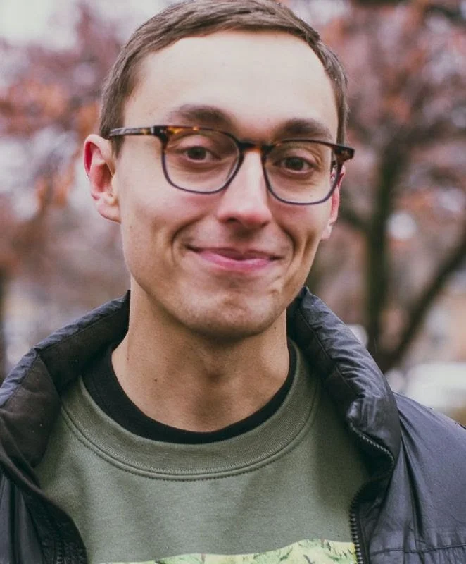 A young man with glasses smiling outdoors, wearing a black jacket over a green shirt, with fall-colored trees in the background.