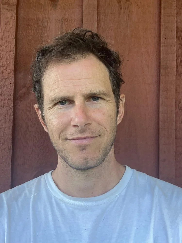 A man with short, curly brown hair and slight stubble, wearing a white T-shirt, standing in front of a red wooden wall.