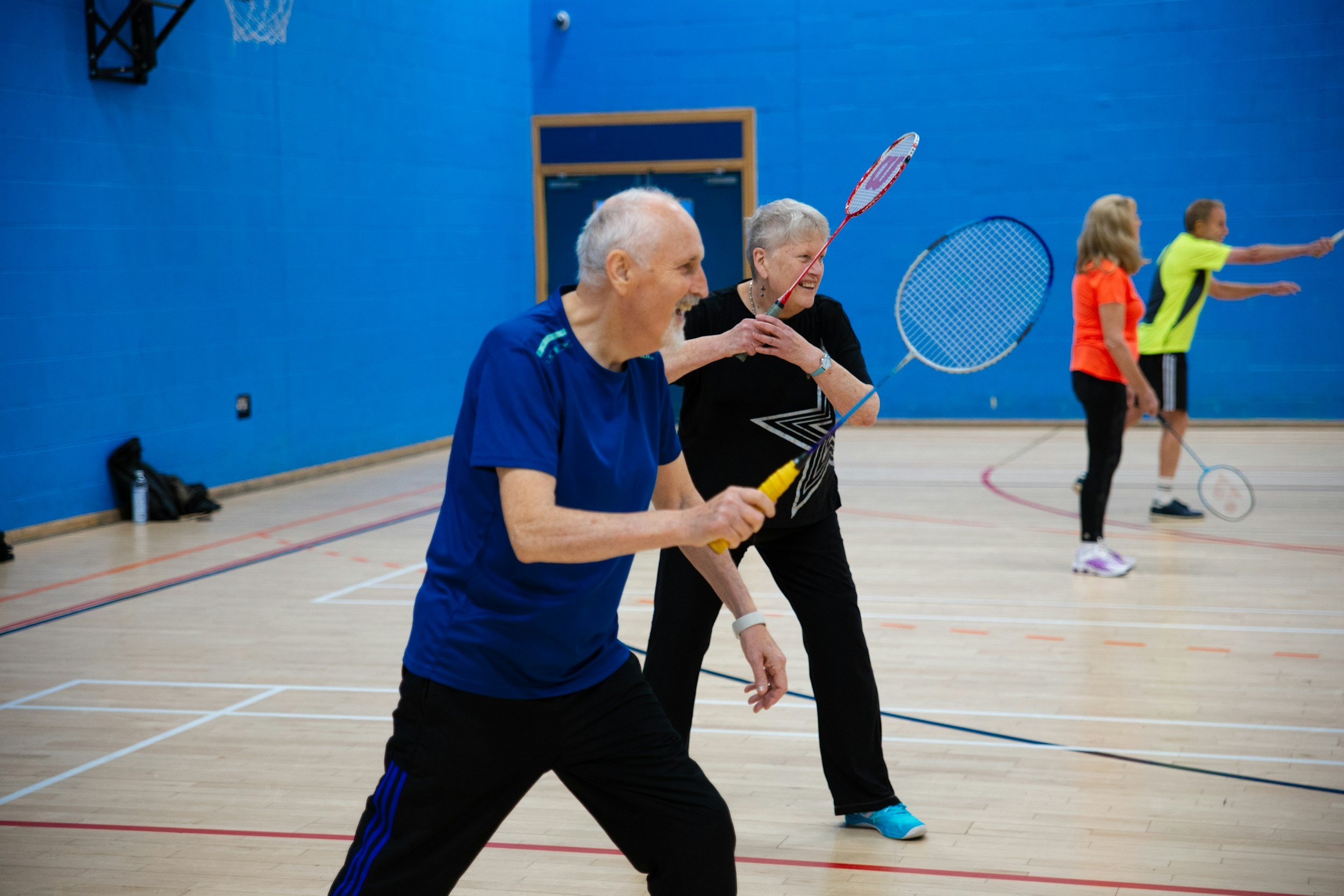 Older man and woman playing badminton indoors, smiling and enjoying the game, highlighting the impact of community involvement for seniors.
