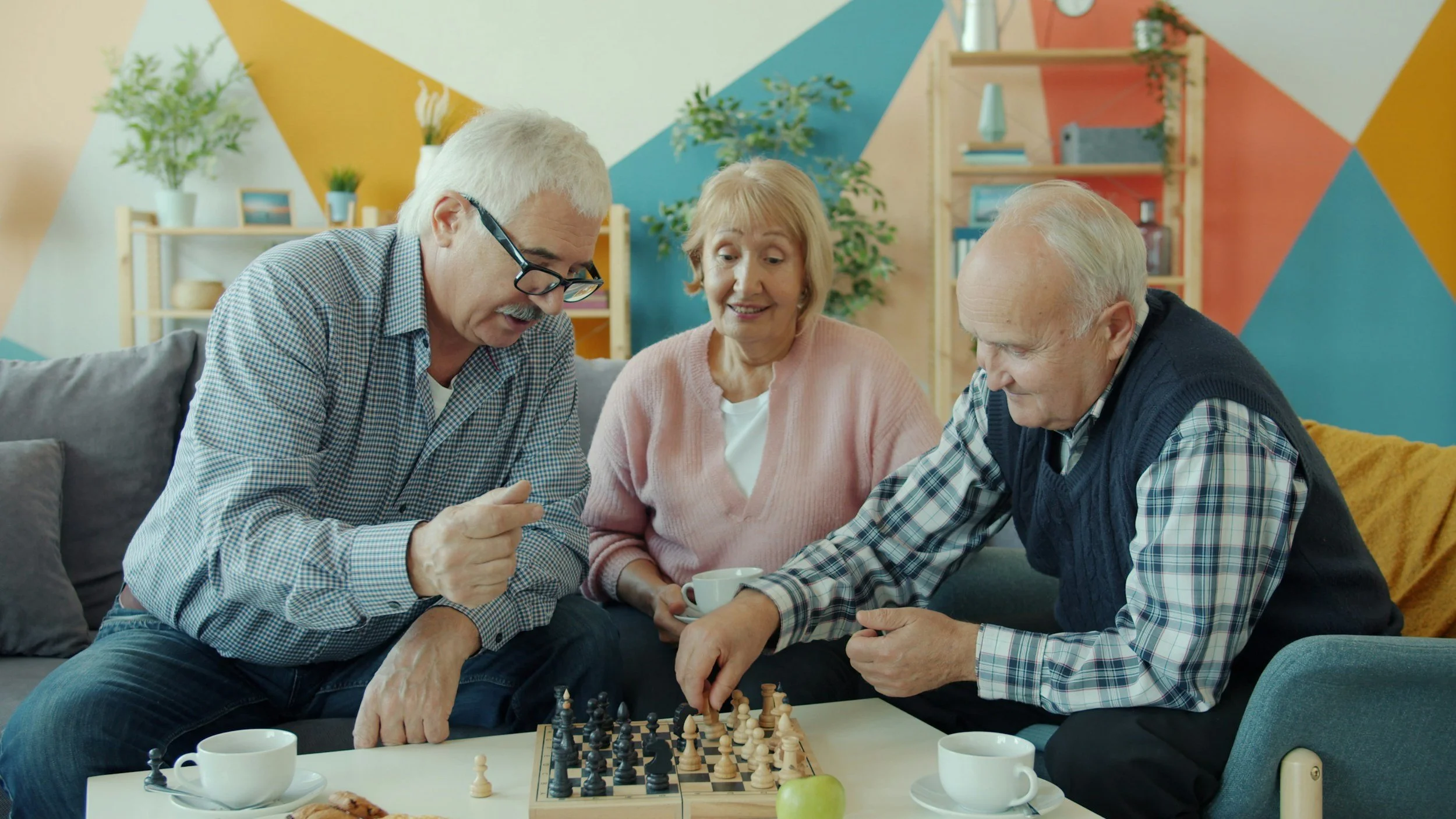 Three elderly people, two men and one woman, playing chess together on a table in a colorful living room.