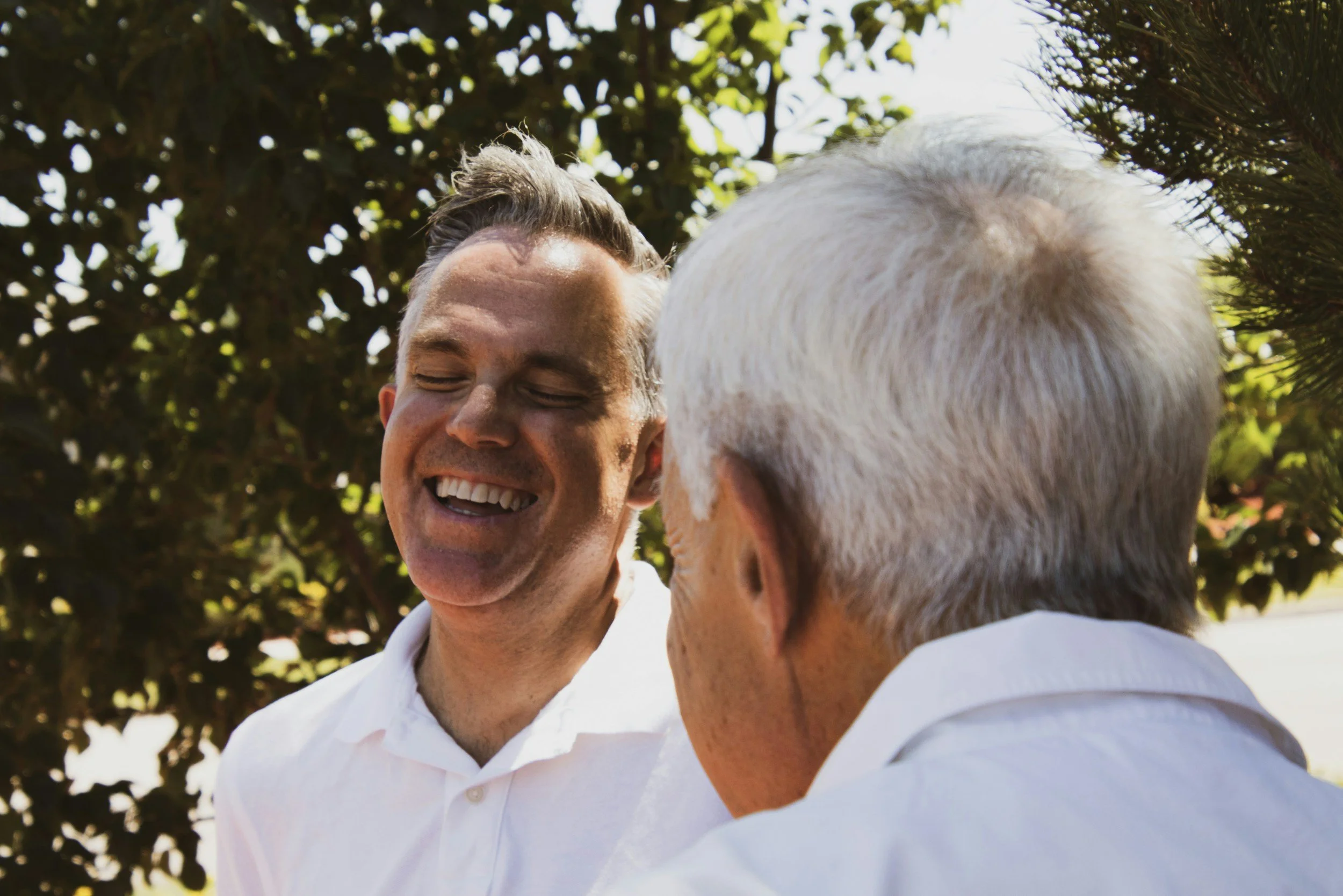 Two men smiling and greeting each other outdoors, with trees in the background.