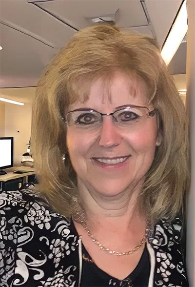 A middle-aged woman with shoulder-length blonde hair and glasses, smiling in an office setting.