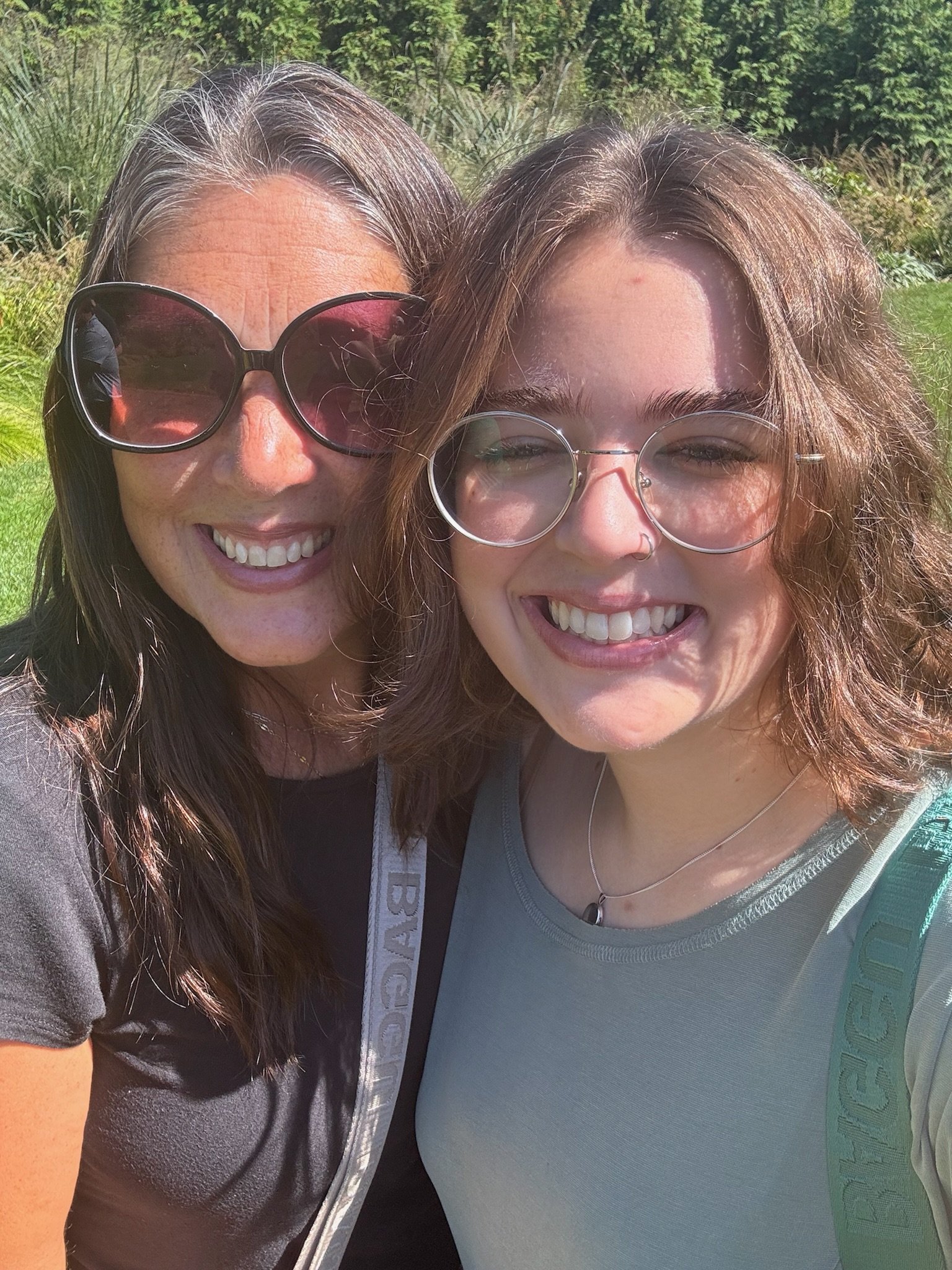 Two women smiling outdoors on a sunny day, with greenery in the background. One is wearing large sunglasses and the other glasses, and they are close together, taking a selfie.