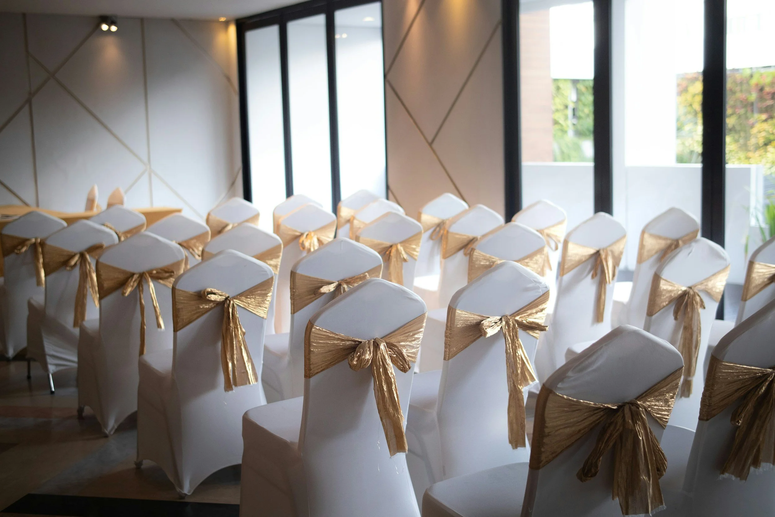 Rows of white chairs with gold sashes tied around the backs, arranged for a formal event in a room with large windows and modern decor.