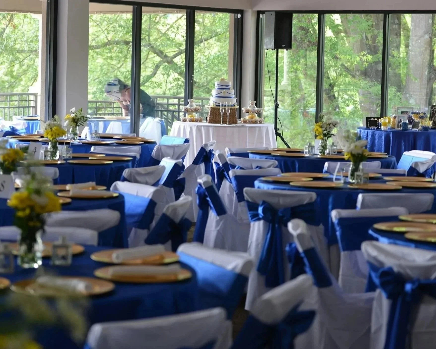 A banquet hall set up for a celebration with round tables covered in blue tablecloths and white chair covers with blue sashes, decorated with yellow and white flower centerpieces, and a dessert table in the background.