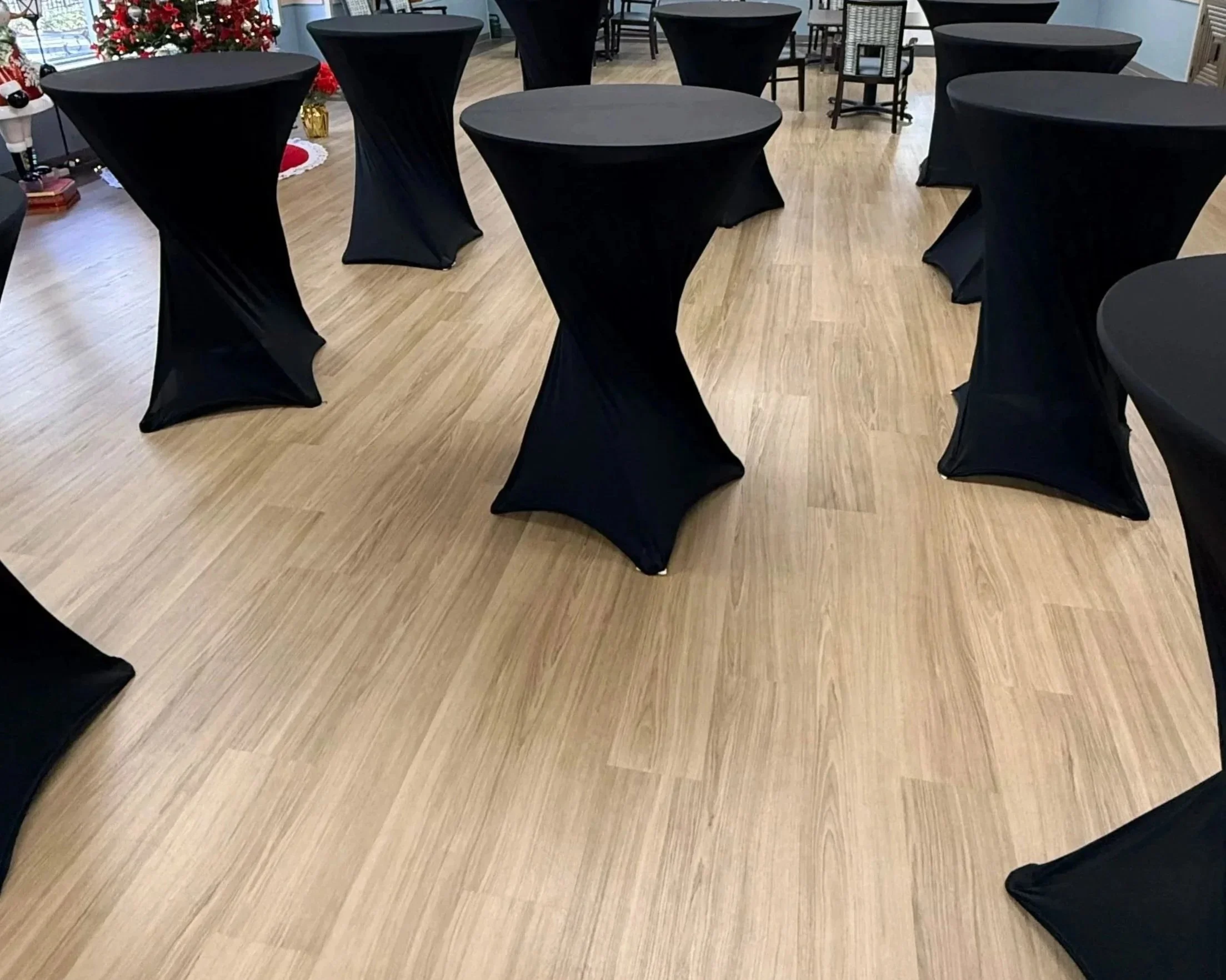 Tall, round cocktail tables covered with black tablecloths are arranged on a wood floor in a room decorated for Christmas.