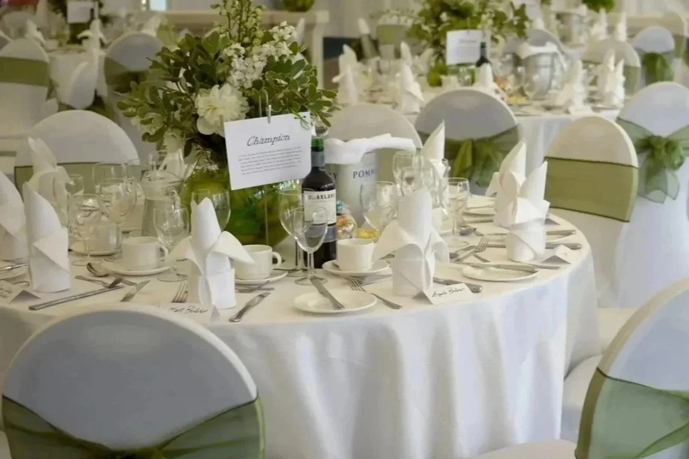 Wedding reception table with white tablecloth, neatly folded white napkins, glassware, and a floral centerpiece with white flowers and greenery.