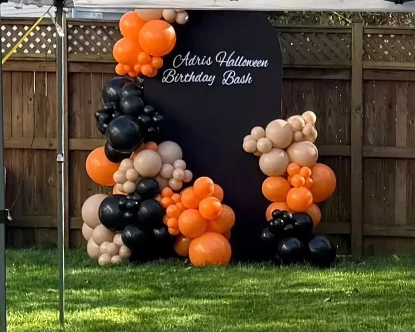Decorative balloon arch with orange, black, and beige balloons in front of a black sign that reads 'Adris Halloween Birthday Bash' on a backyard grass lawn with a wooden fence in the background.